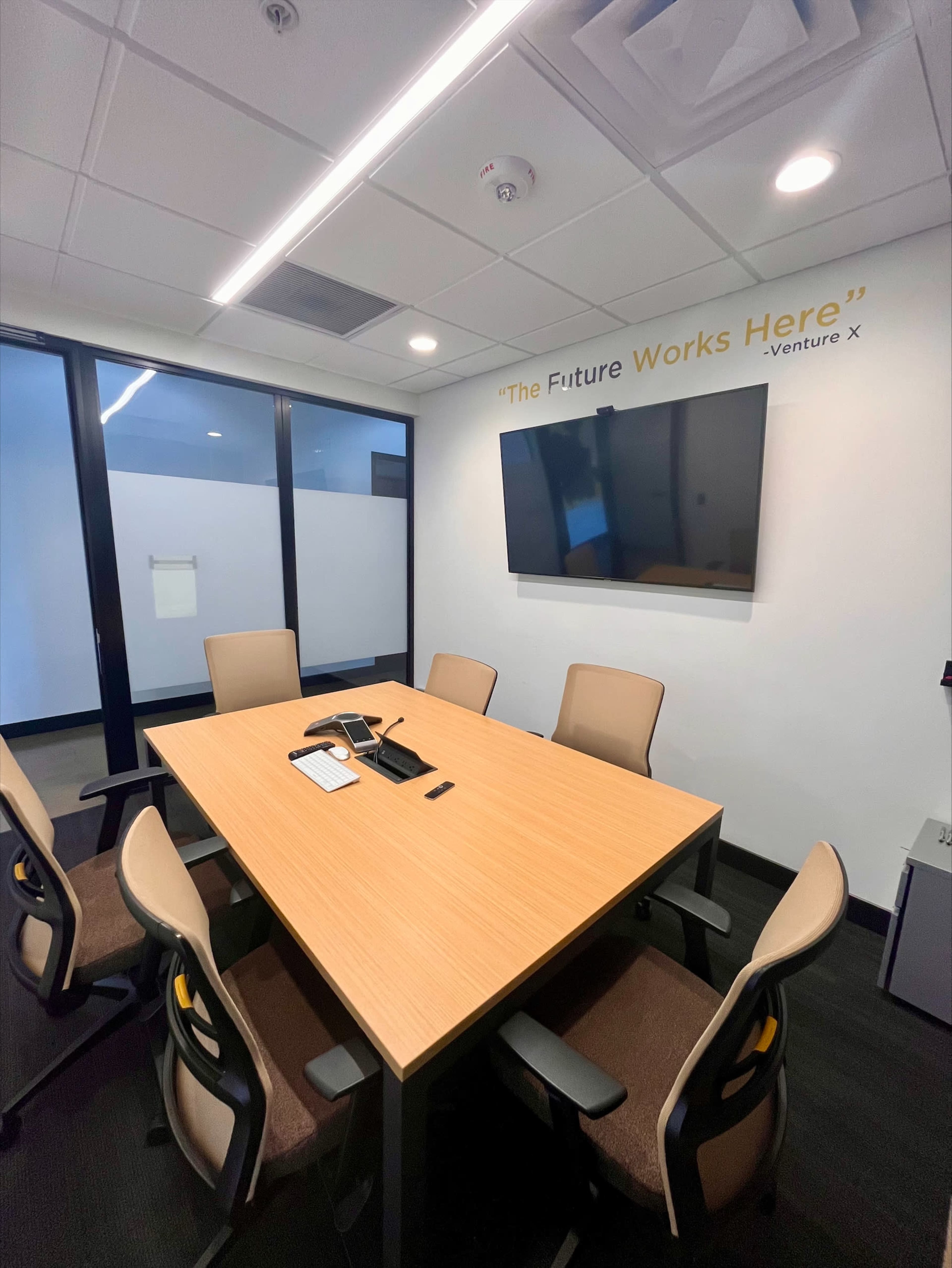 A modern conference room with a wooden table, six chairs, a large TV mounted on the wall, and a motivational quote above the screen.