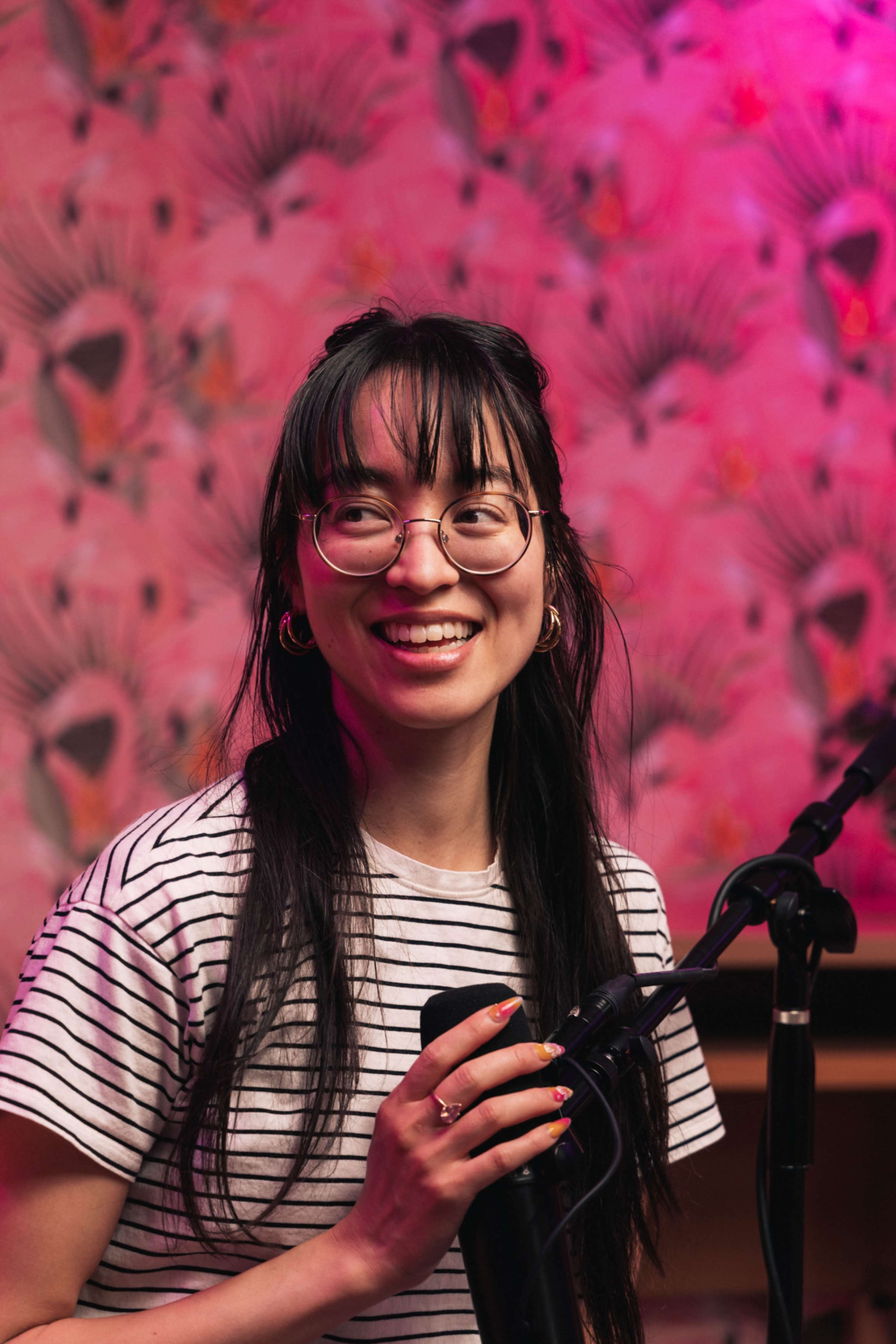 A young woman with long black hair and glasses stands in front of a microphone against a brightly patterned pink wall.