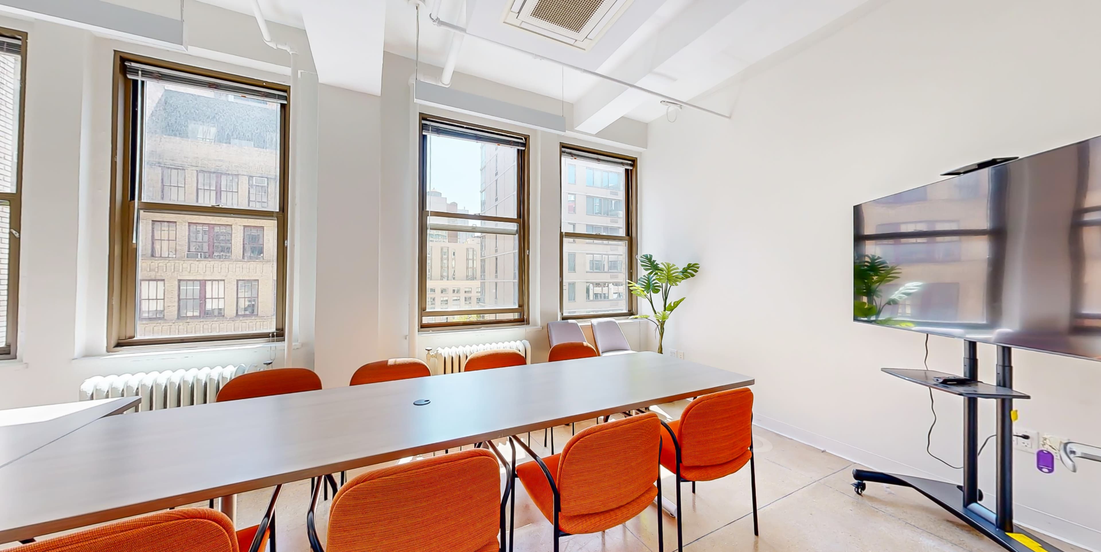 A modern conference room features a long table surrounded by orange chairs, with large windows allowing natural light to fill the space.
