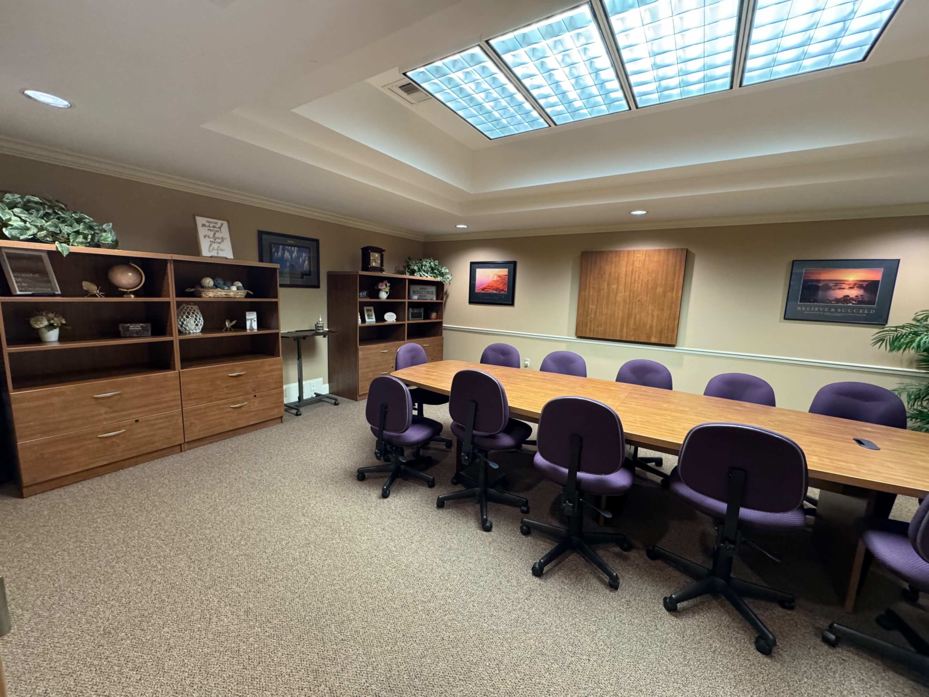 A conference room features a long wooden table surrounded by purple office chairs, with shelving units and framed artwork on the walls.