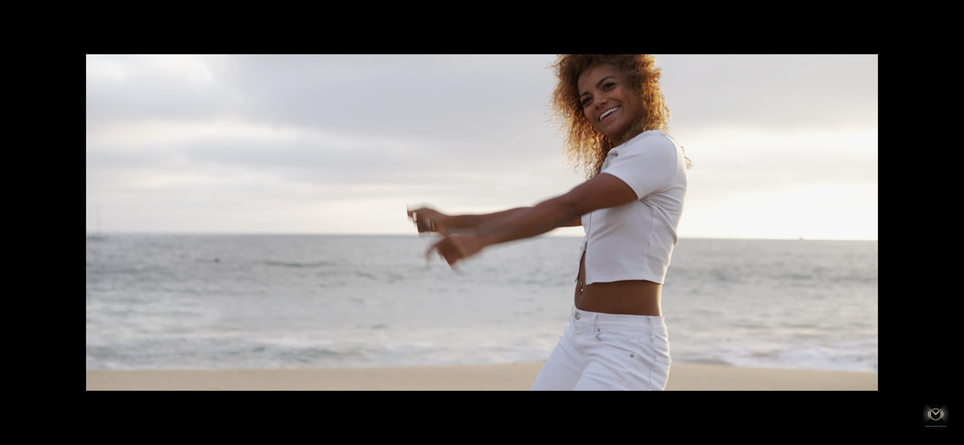 A person in a white outfit dances near the shoreline with the ocean in the background.