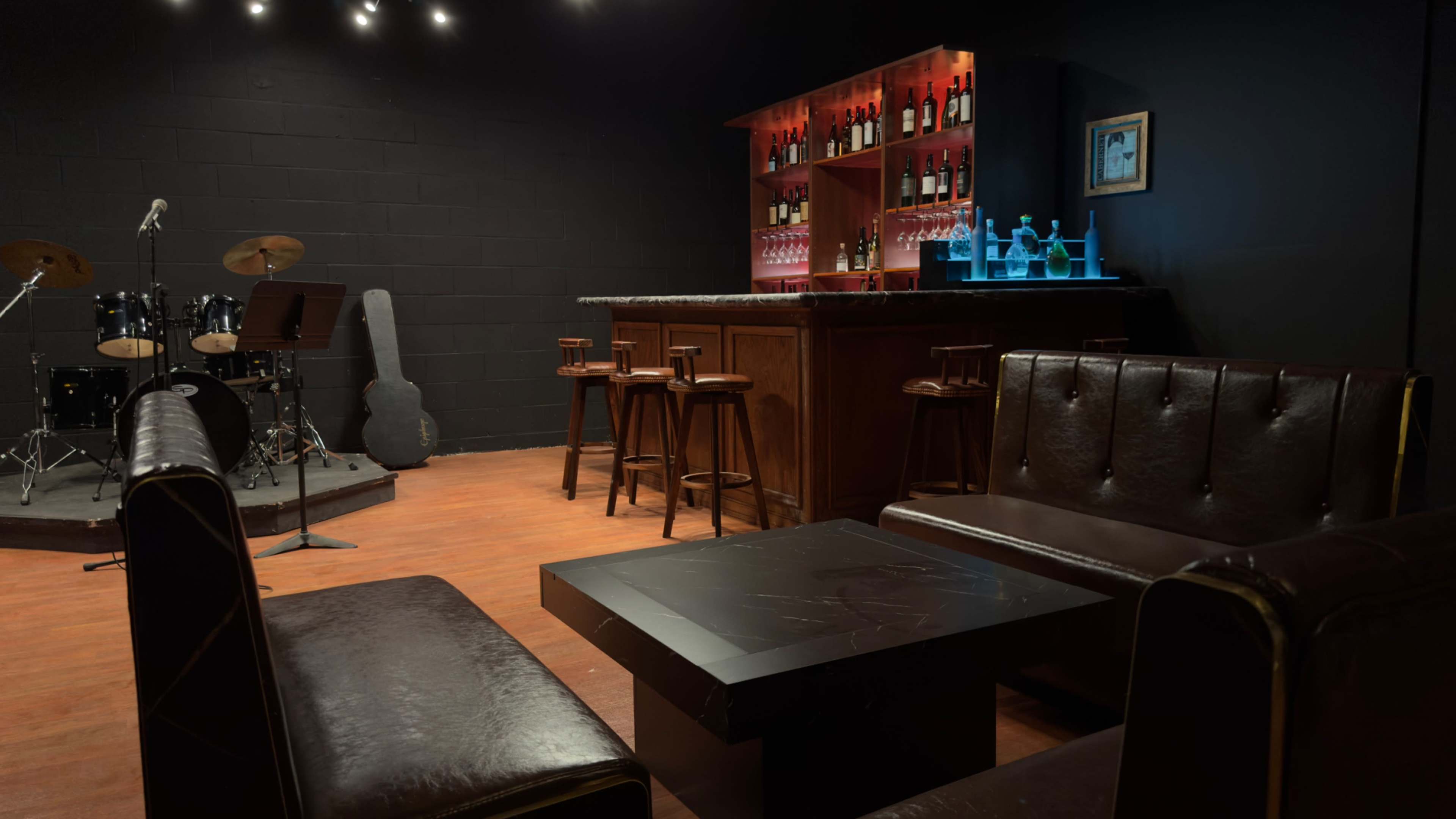 The scene shows a dimly lit bar area with a stage for live music, featuring a wooden bar stocked with drinks, a few high stools, and leather seating arrangements.