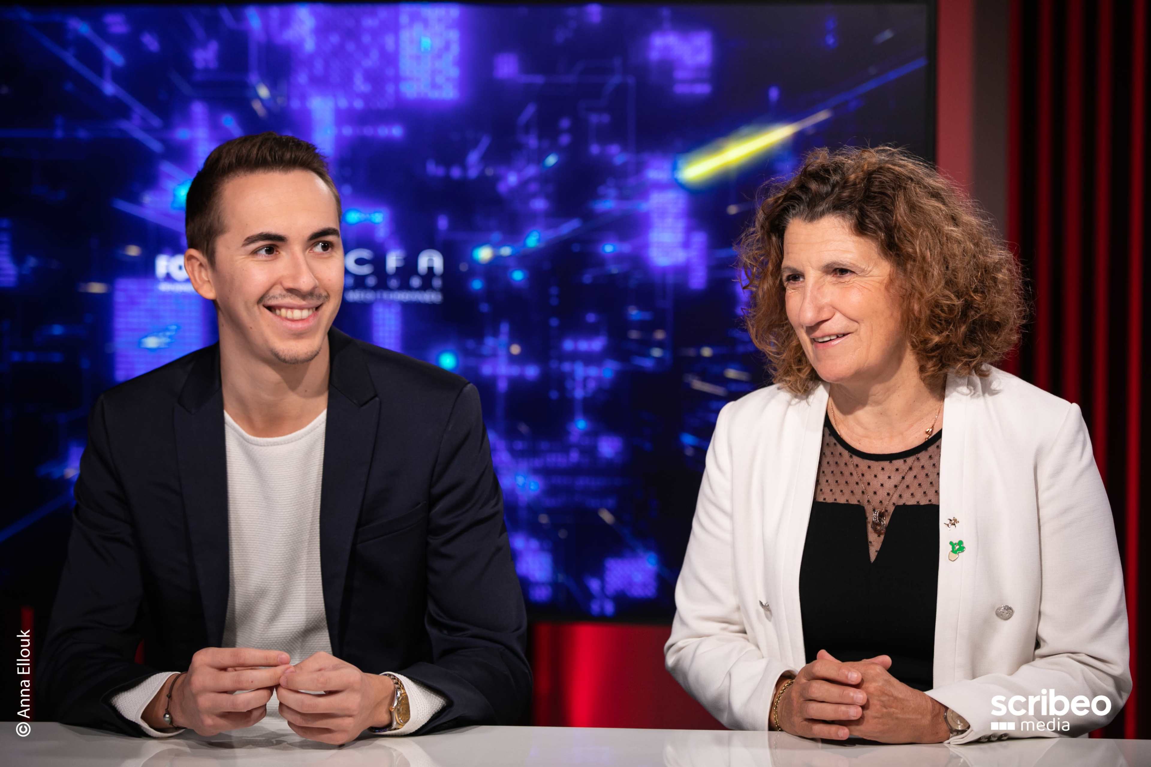 A young man and a woman sit at a table in a studio, facing the camera with a digital backdrop.