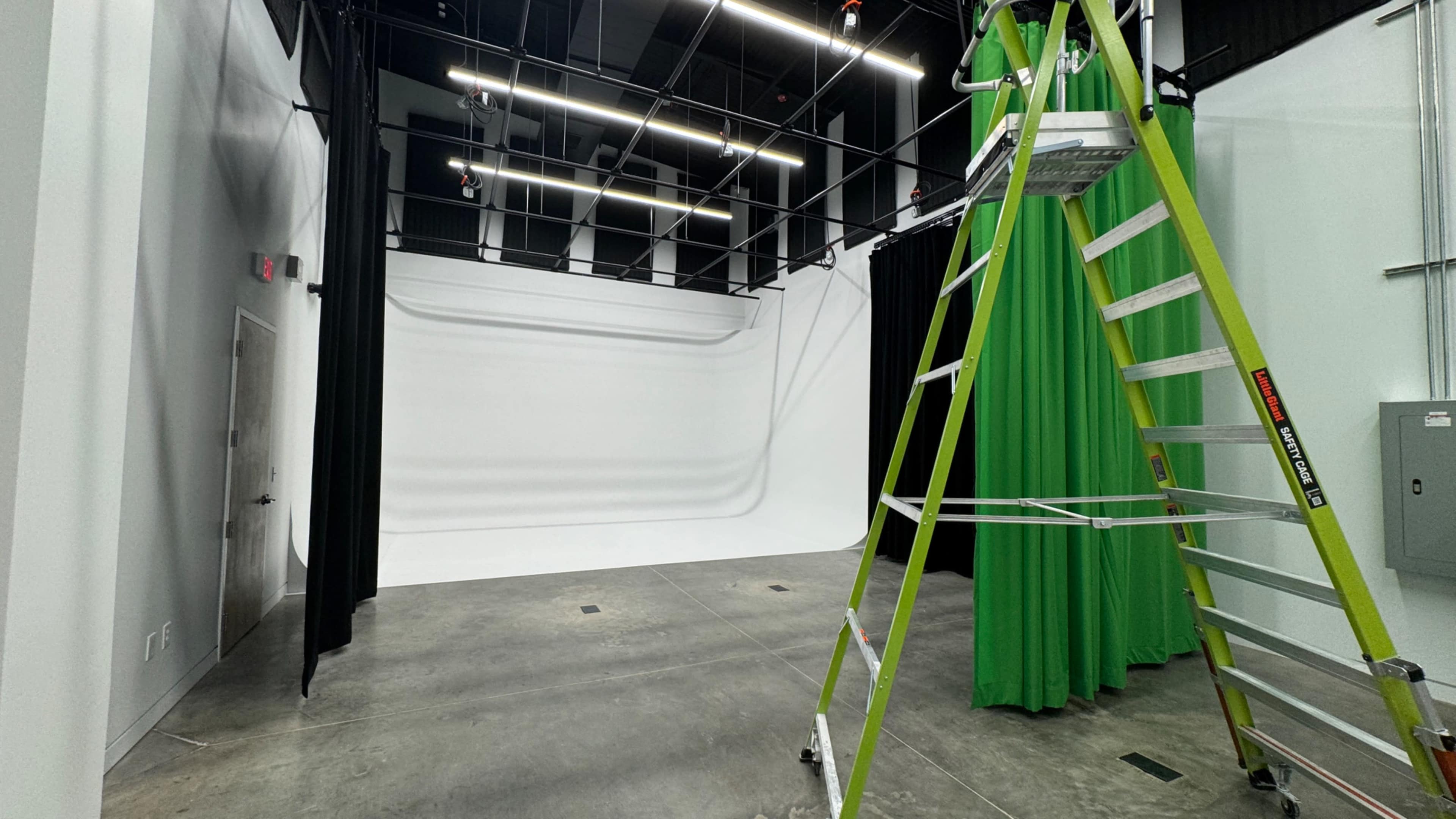 The image shows an empty studio space featuring a white backdrop, black curtains, and a green screen, alongside a folding ladder.