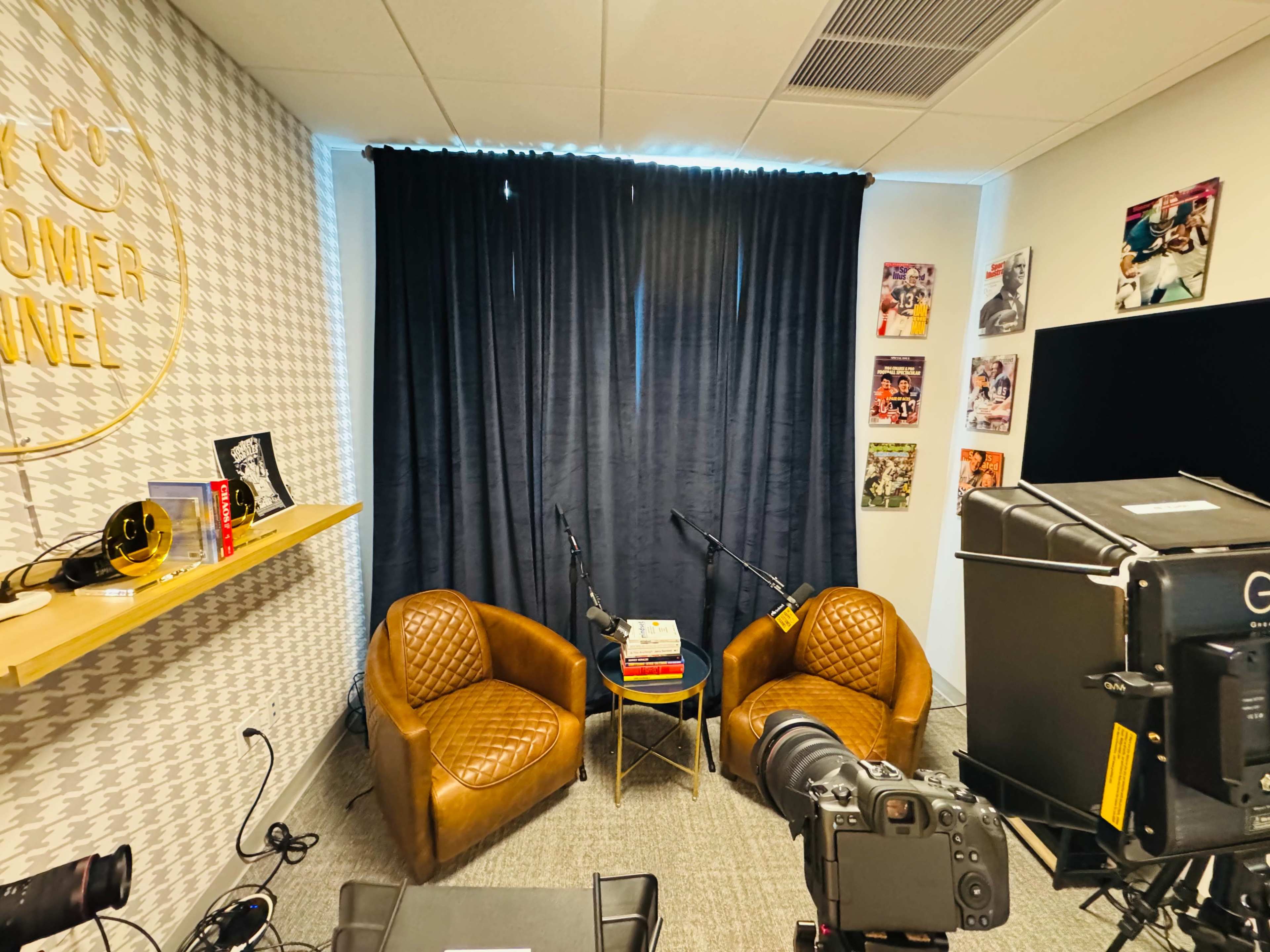 The image shows a small podcast recording studio with two brown chairs, a table stacked with books, and microphones on stands against a backdrop of a black curtain and wall decorations.