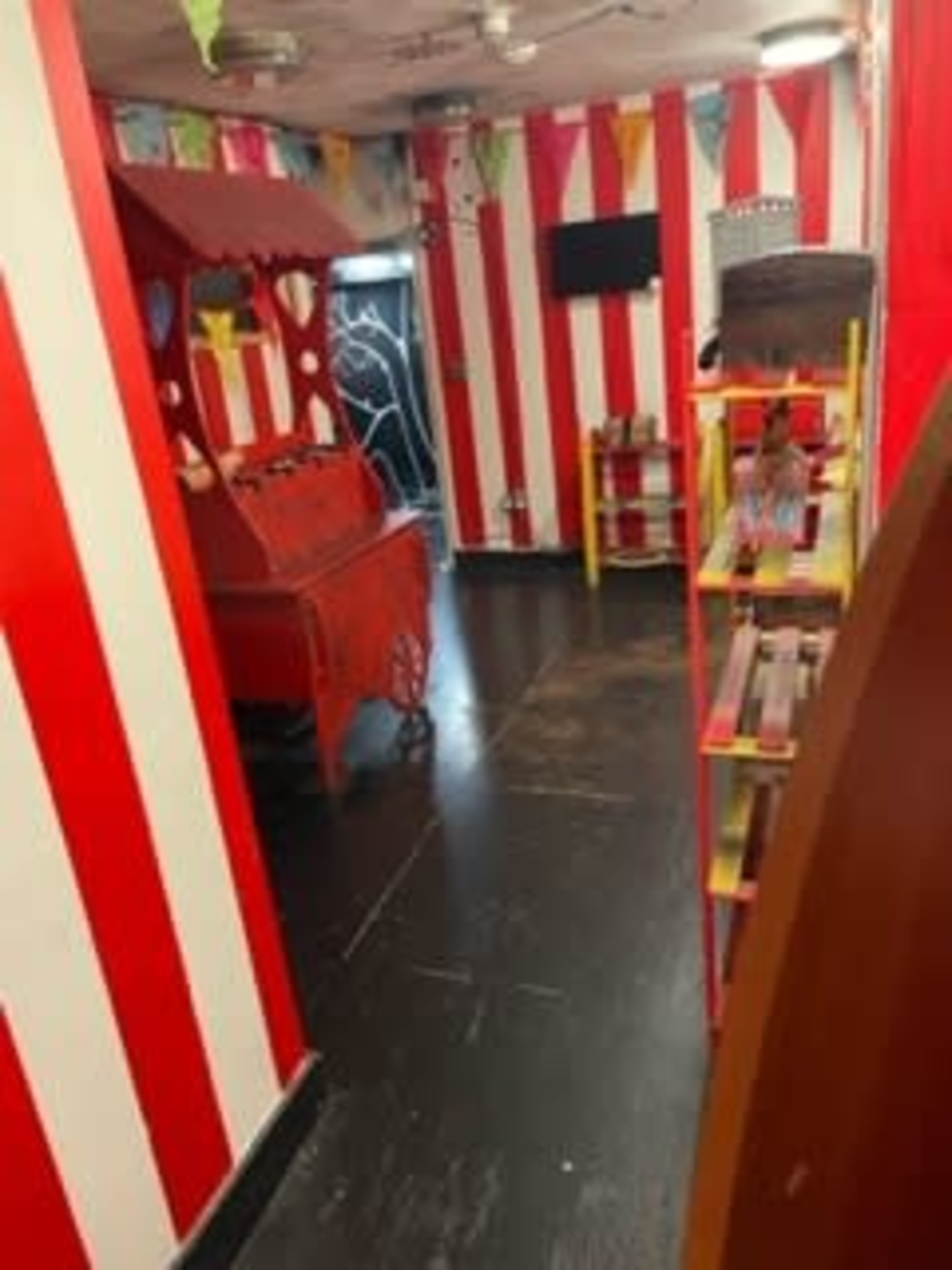 A narrow hallway decorated with red and white striped walls, featuring a carnival-themed booth on one side and a shelf displaying various items on the other.
