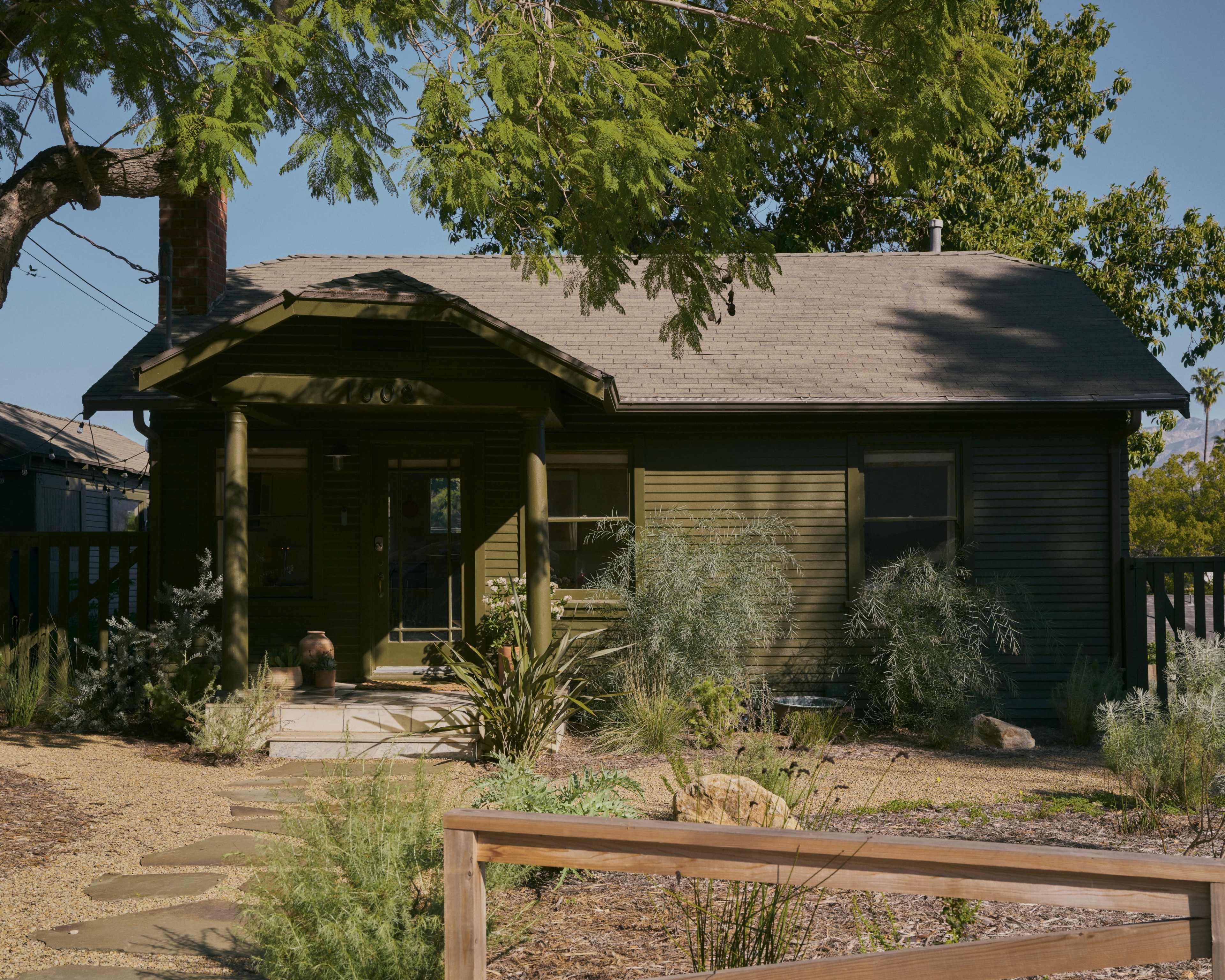 The image shows a green, single-story house surrounded by a landscaped yard with various plants and a stone pathway leading to the entrance.