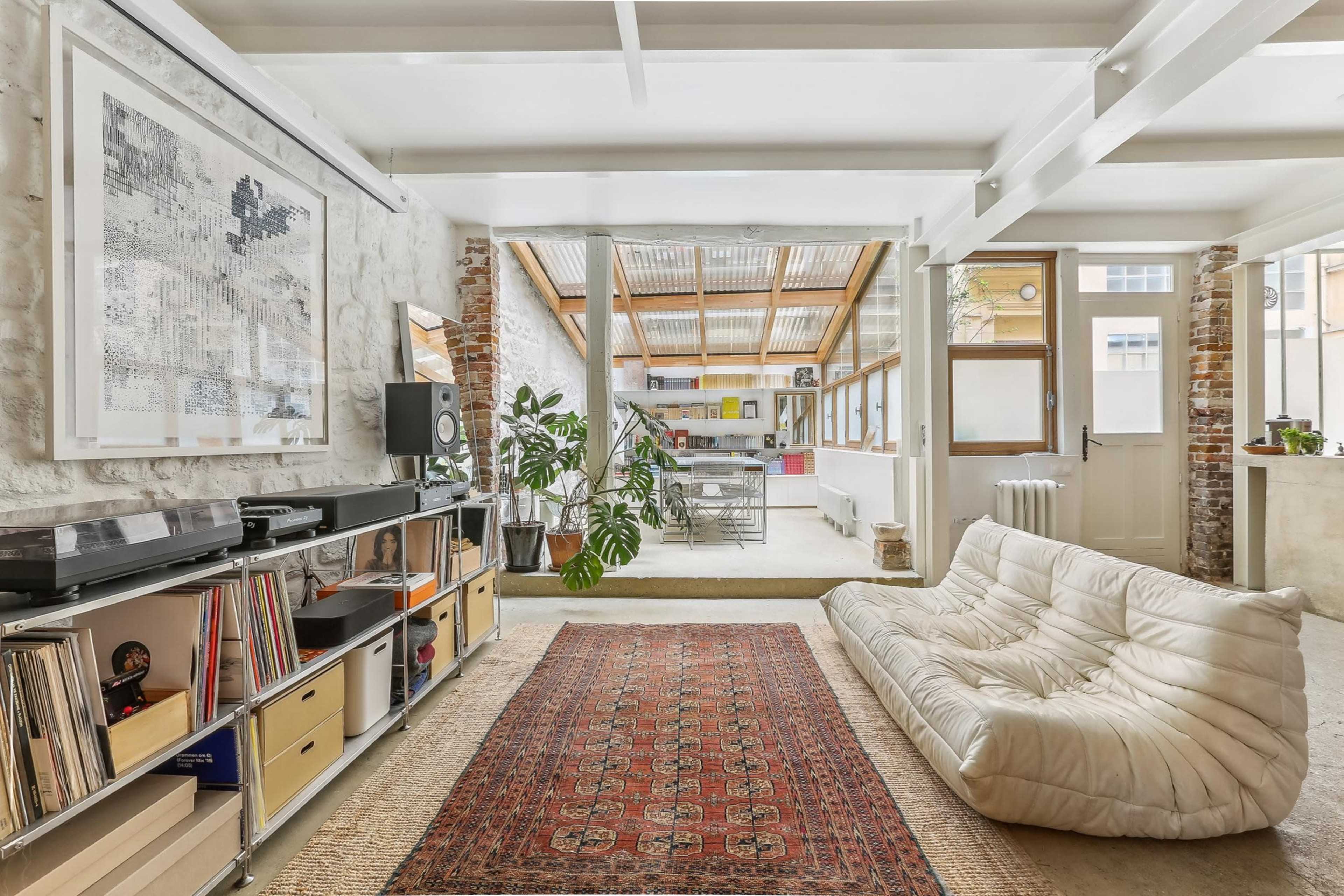 The image shows a modern, open-concept living space featuring a plush sofa, a record player setup, a colorful area rug, and large windows that let in natural light.