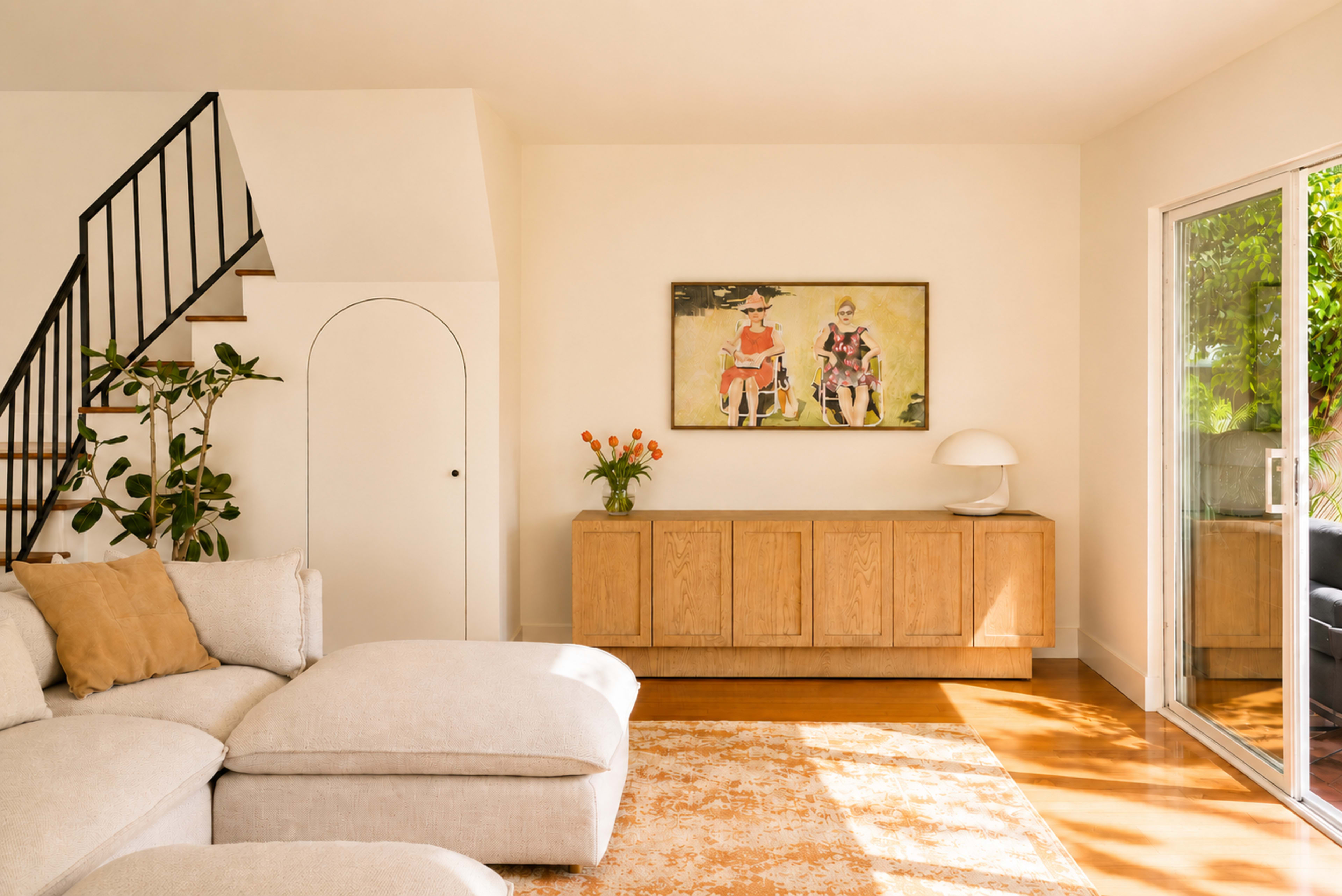 The image shows a modern living room with a light-colored sectional sofa, a wooden sideboard, an art piece depicting two seated figures, and a large glass door opening to a patio.