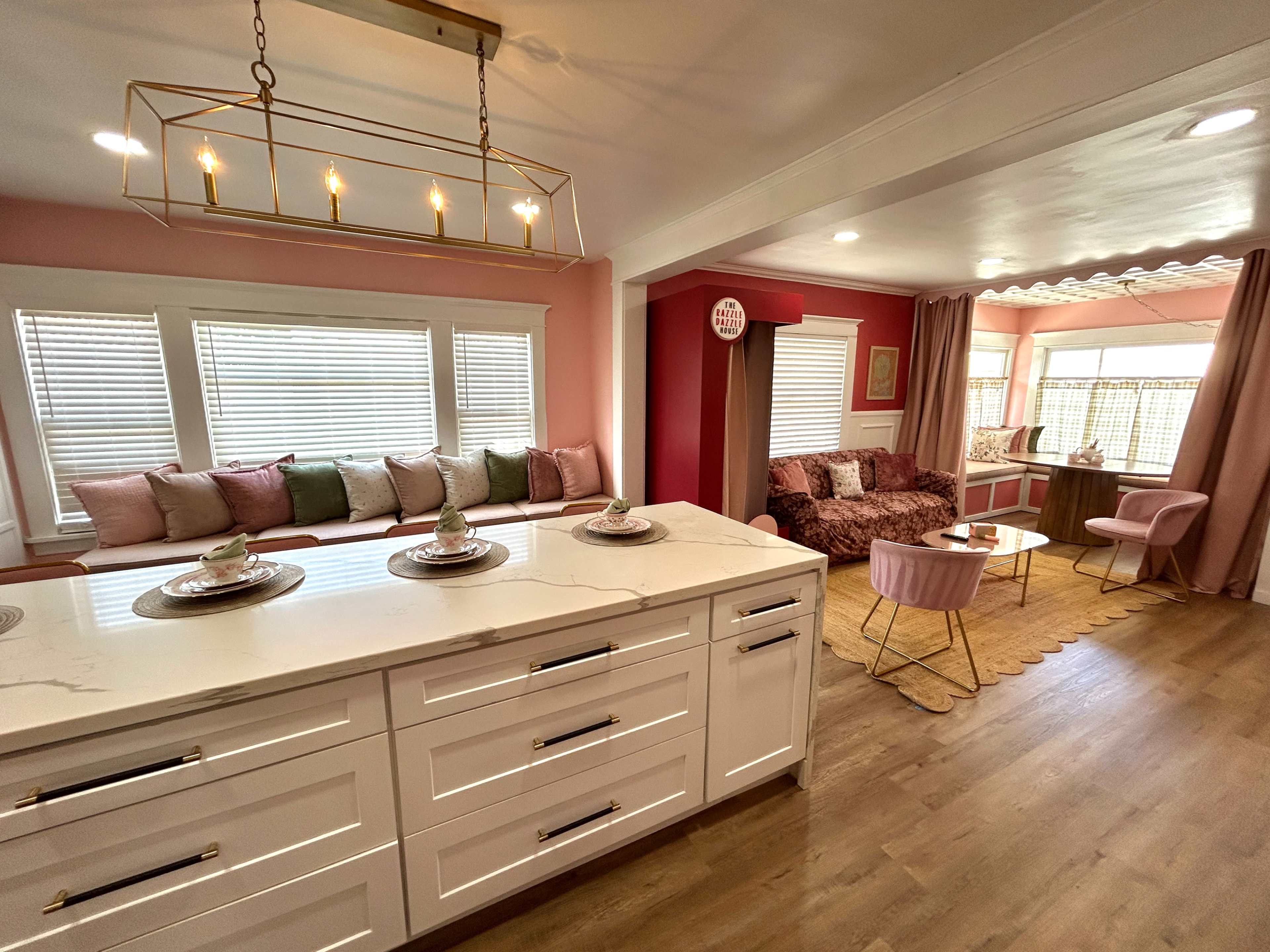 The image shows a modern kitchen opening into a cozy living area, featuring pink walls, a marble island with decorative plates, and comfortable seating.