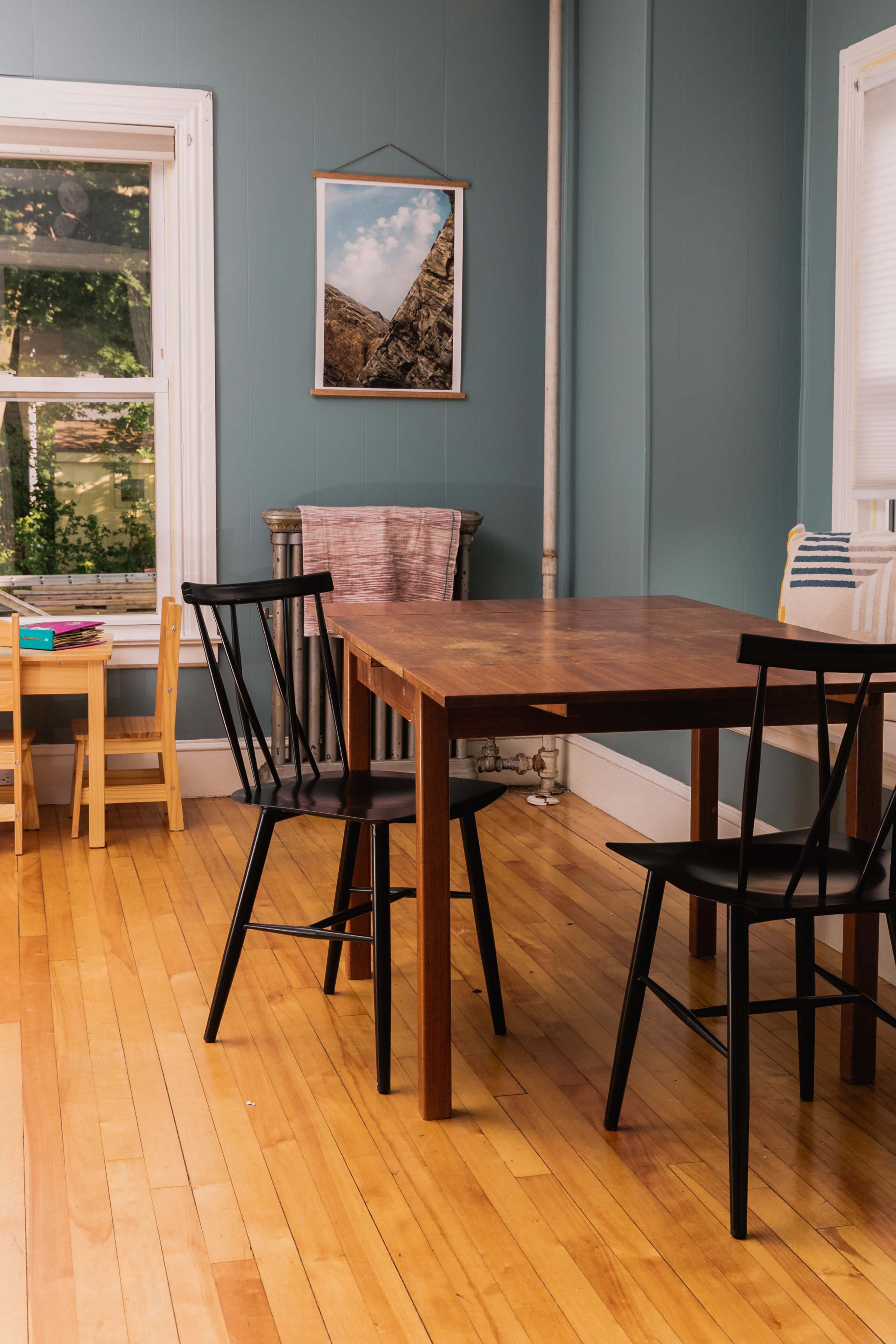 A wooden table with black chairs is situated in a room with blue walls, featuring a large window and a framed photograph on the wall.