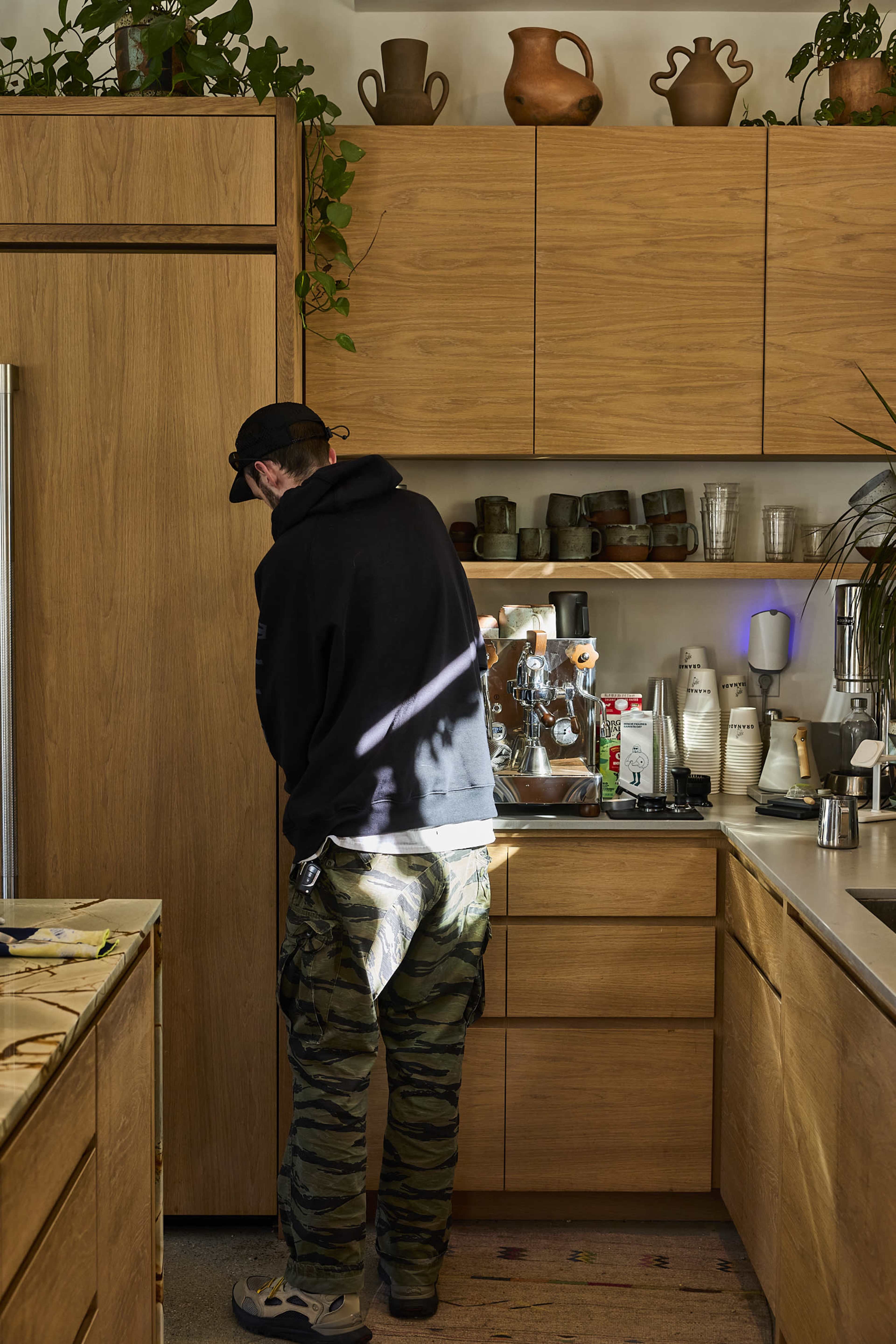 A person in a black hoodie and camouflage pants stands in a modern kitchen, leaning into a wooden cabinet while surrounded by various kitchen items and plants.