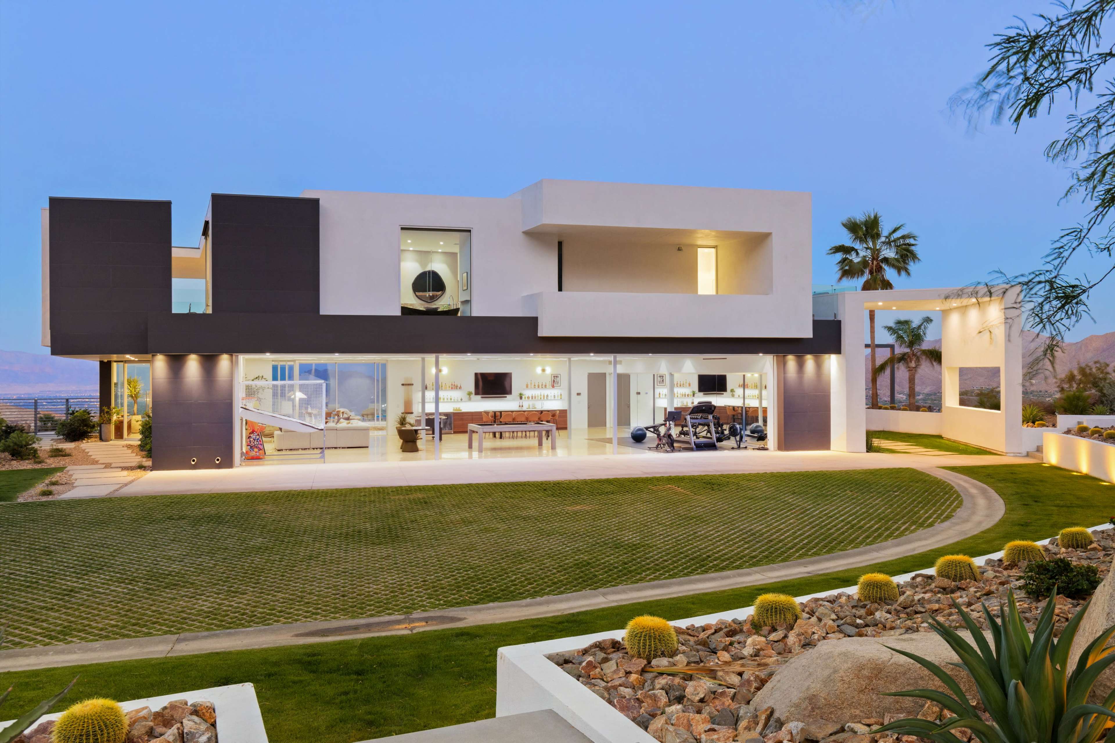 A modern two-story house with a flat roof features a large lawn, patio, and panoramic windows overlooking the landscape.
