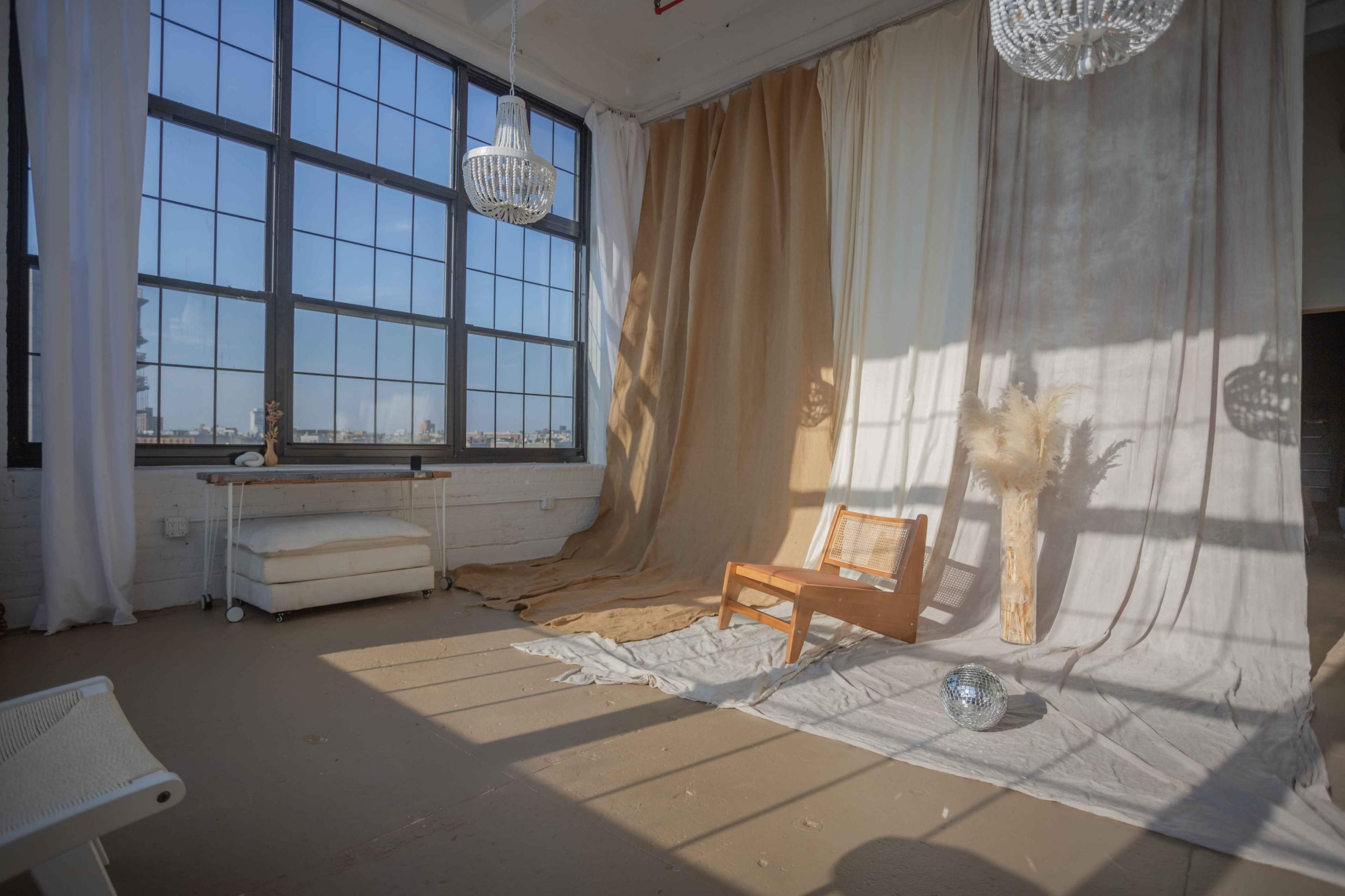 The scene features a spacious room with large windows, draped fabric backdrops, a wooden chair, and decorative elements like a floor vase and a disco ball.
