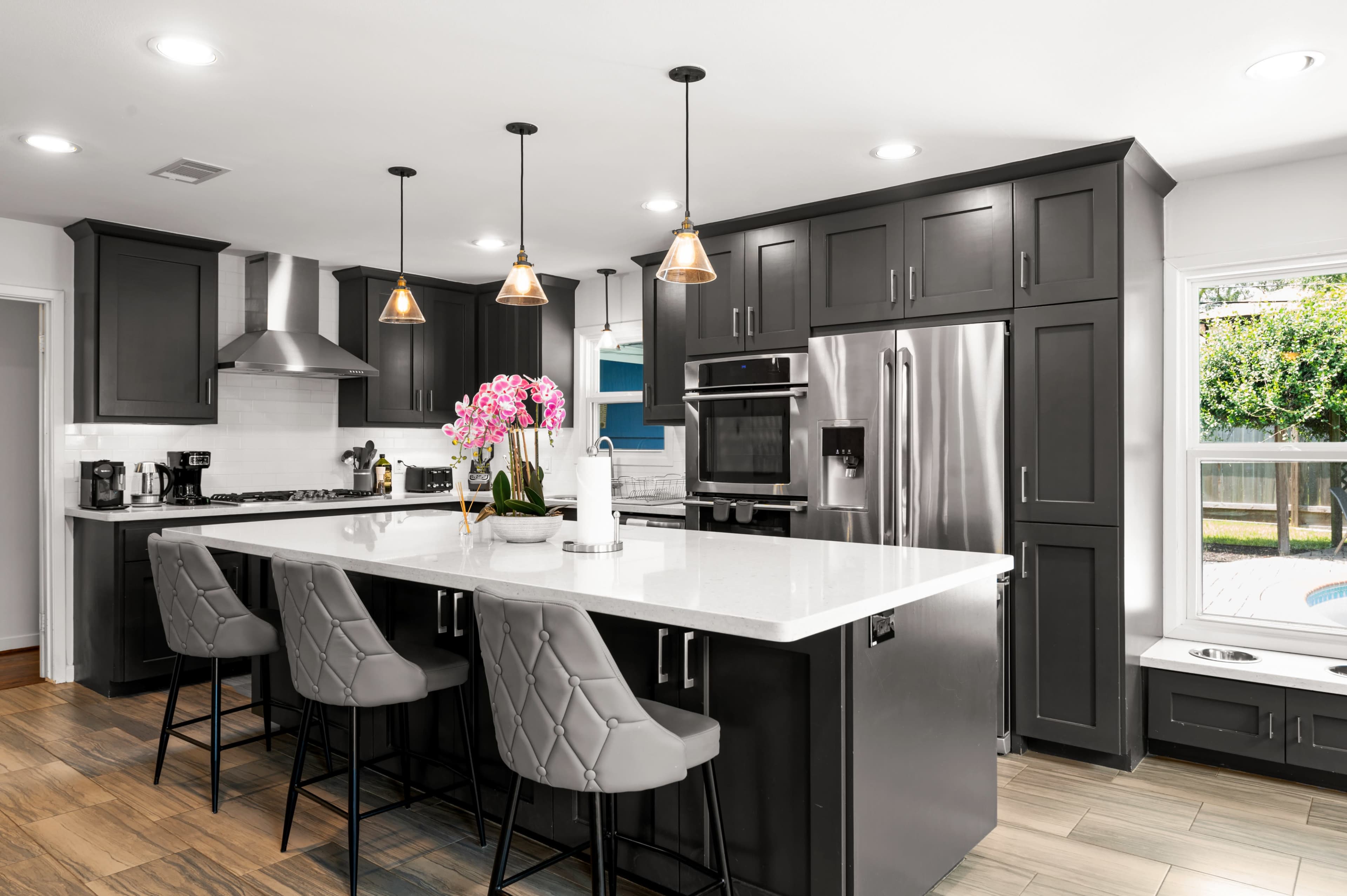 A modern kitchen with dark cabinetry, stainless steel appliances, a large white countertop island with seating, and a vase of pink orchids.