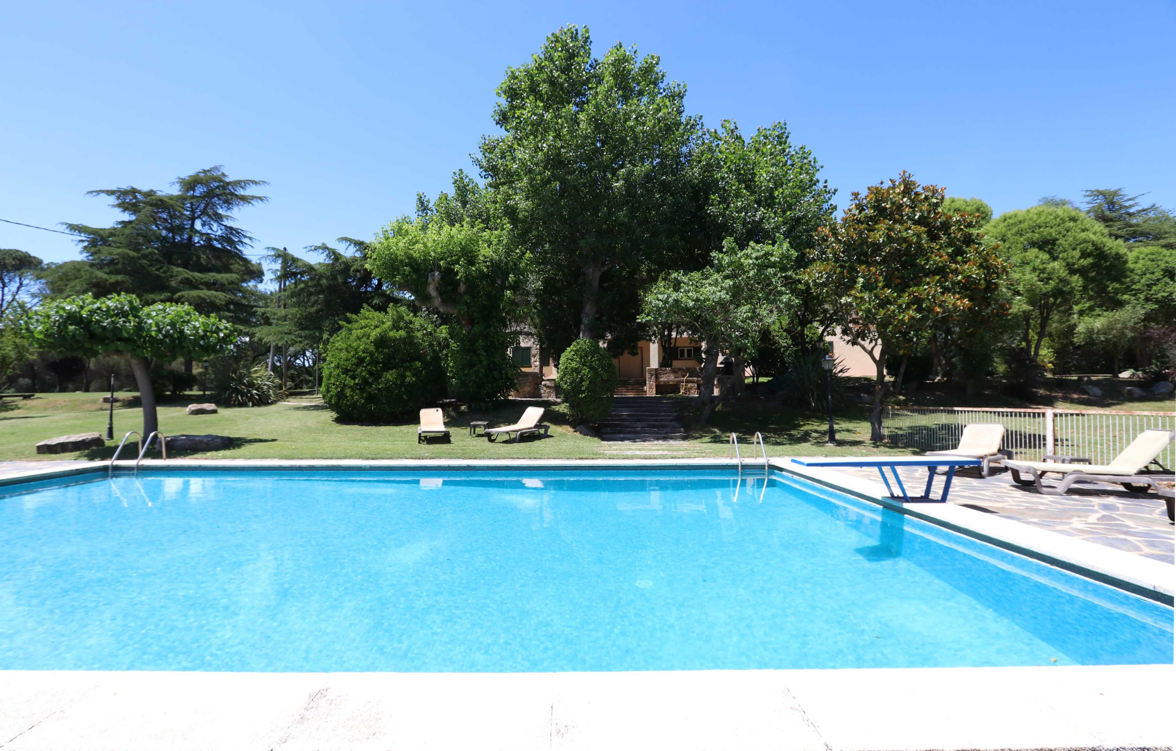 A clear swimming pool is surrounded by a grassy area and trees, with a house visible in the background.