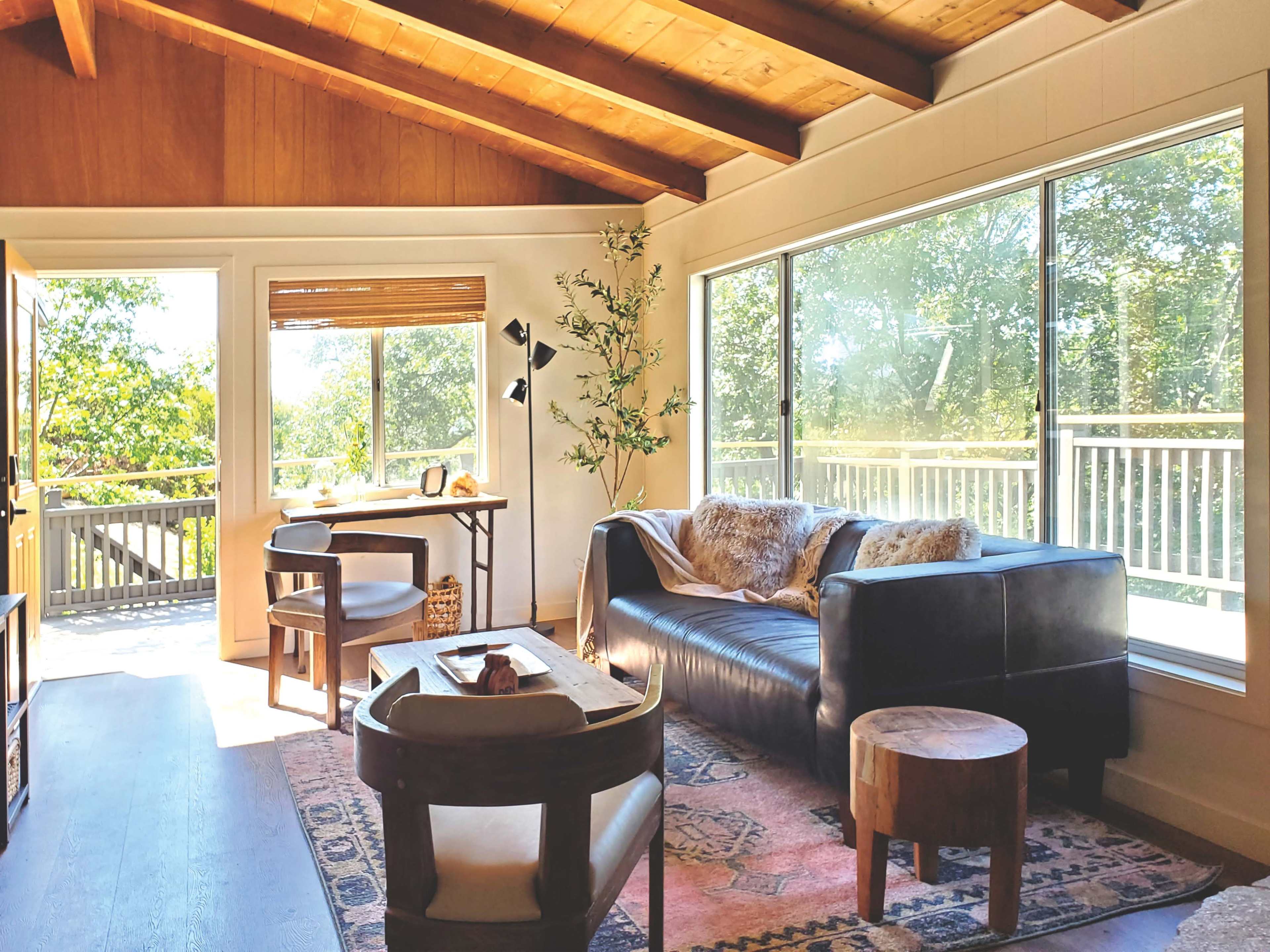 A cozy living room features a dark sofa, a wooden table, and large windows that overlook a tree-filled outdoor space.