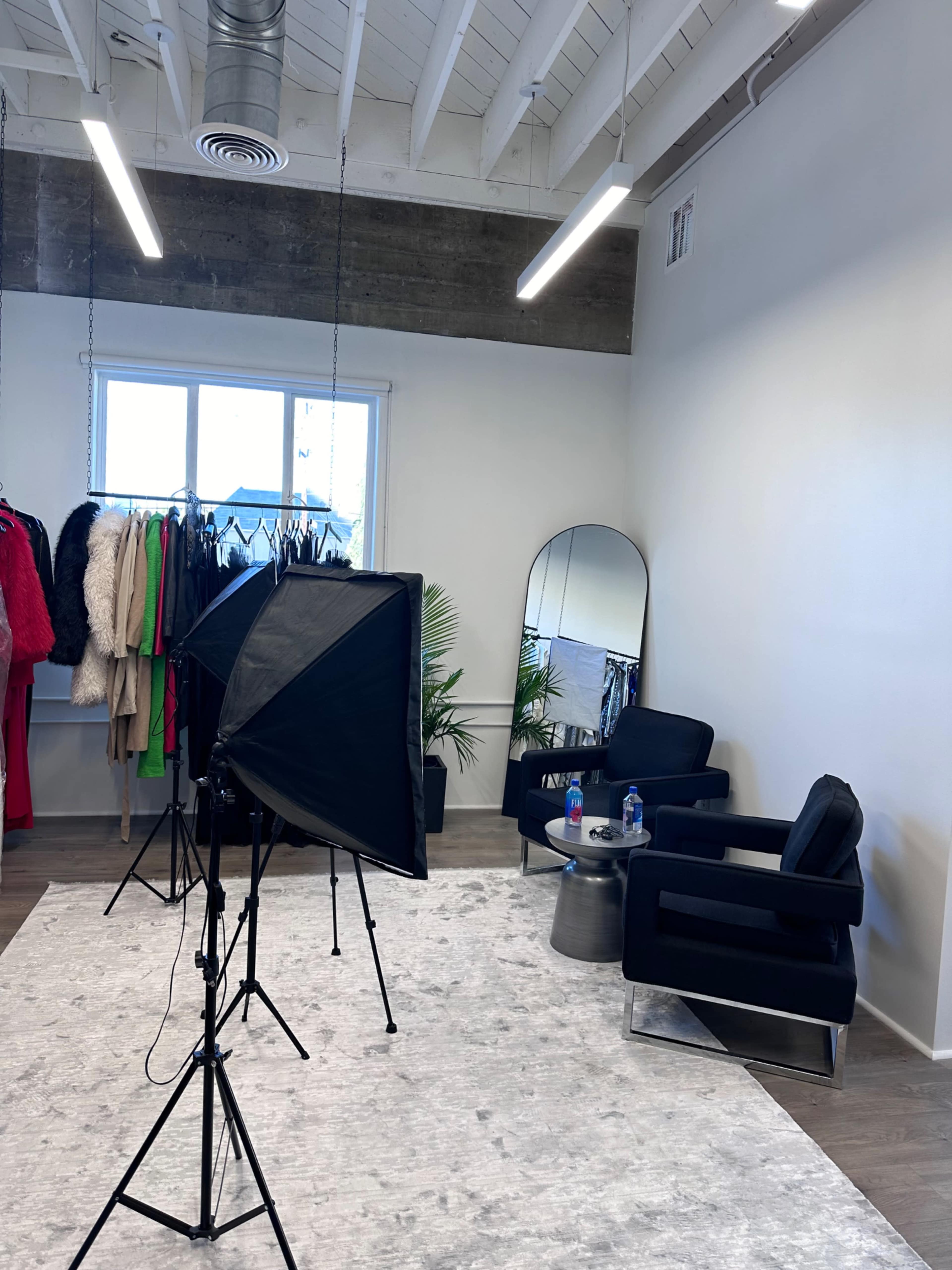 A photography setup inside a spacious room featuring a clothing rack, a full-length mirror, and two black chairs on light-colored flooring.