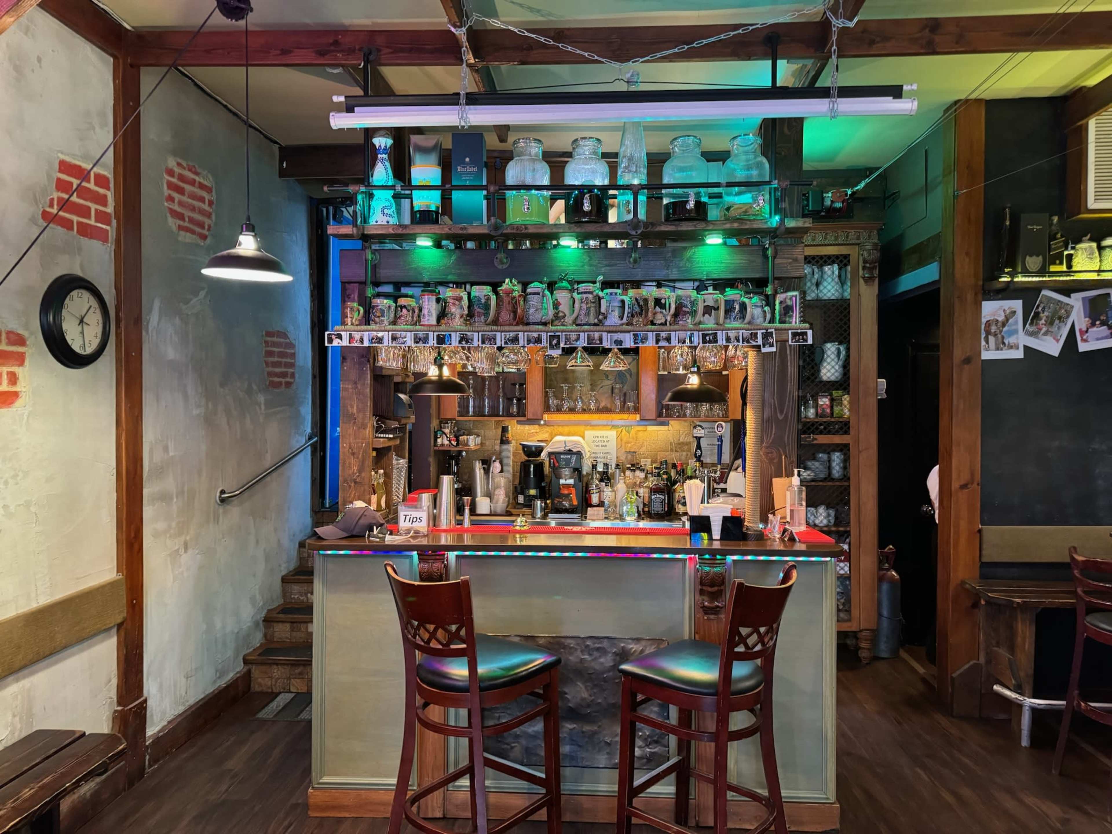 The image shows a cozy bar interior with a countertop, chairs, and shelves displaying jars and decor under colorful lighting.