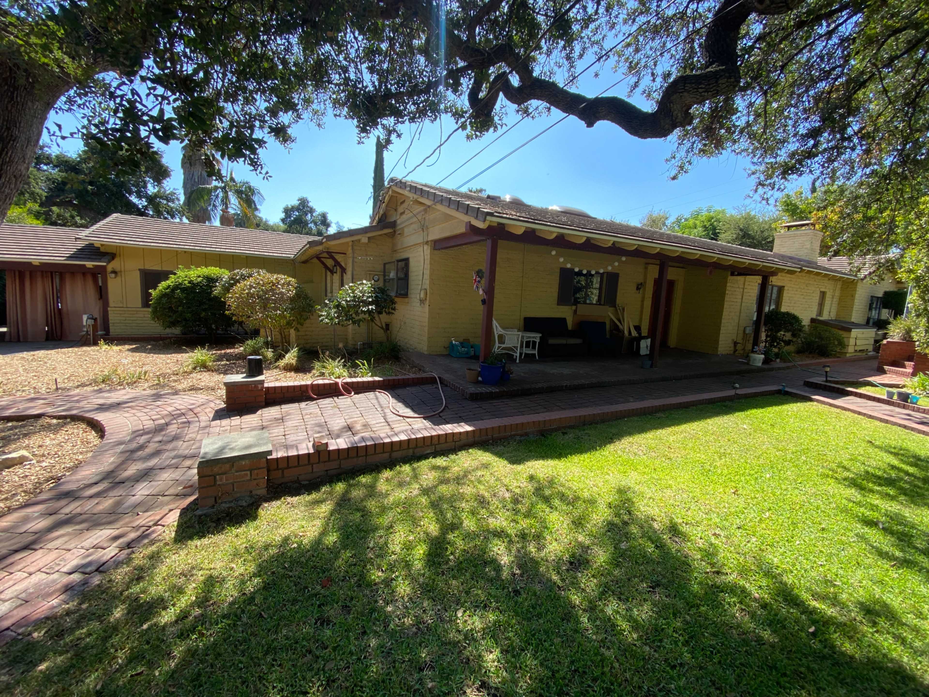 A yellow brick house with a porch surrounded by a lawn, trees, and a brick walkway.