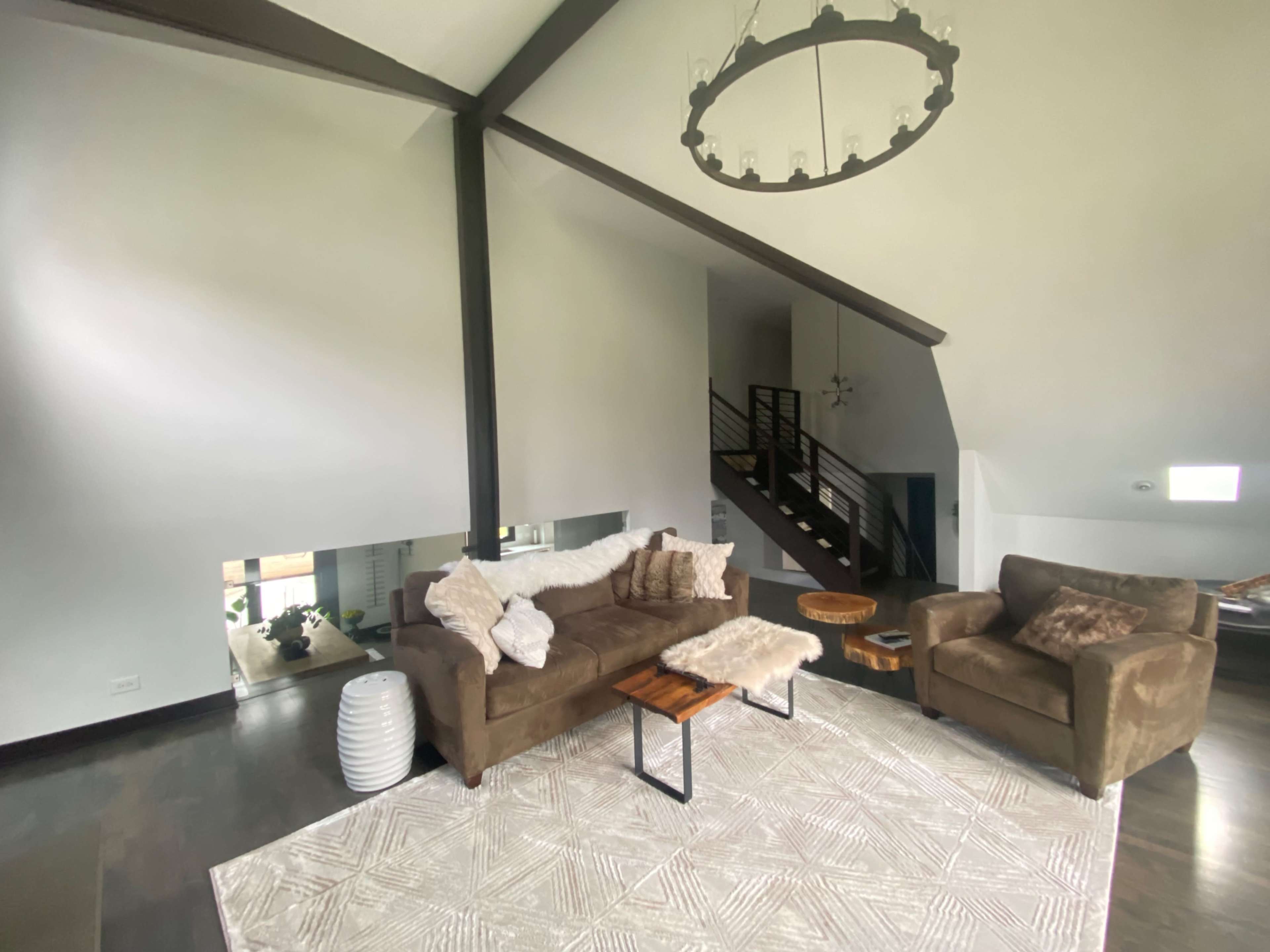 A spacious living room features a brown sectional sofa, a light area rug, and wooden accent tables, with a staircase visible in the background.