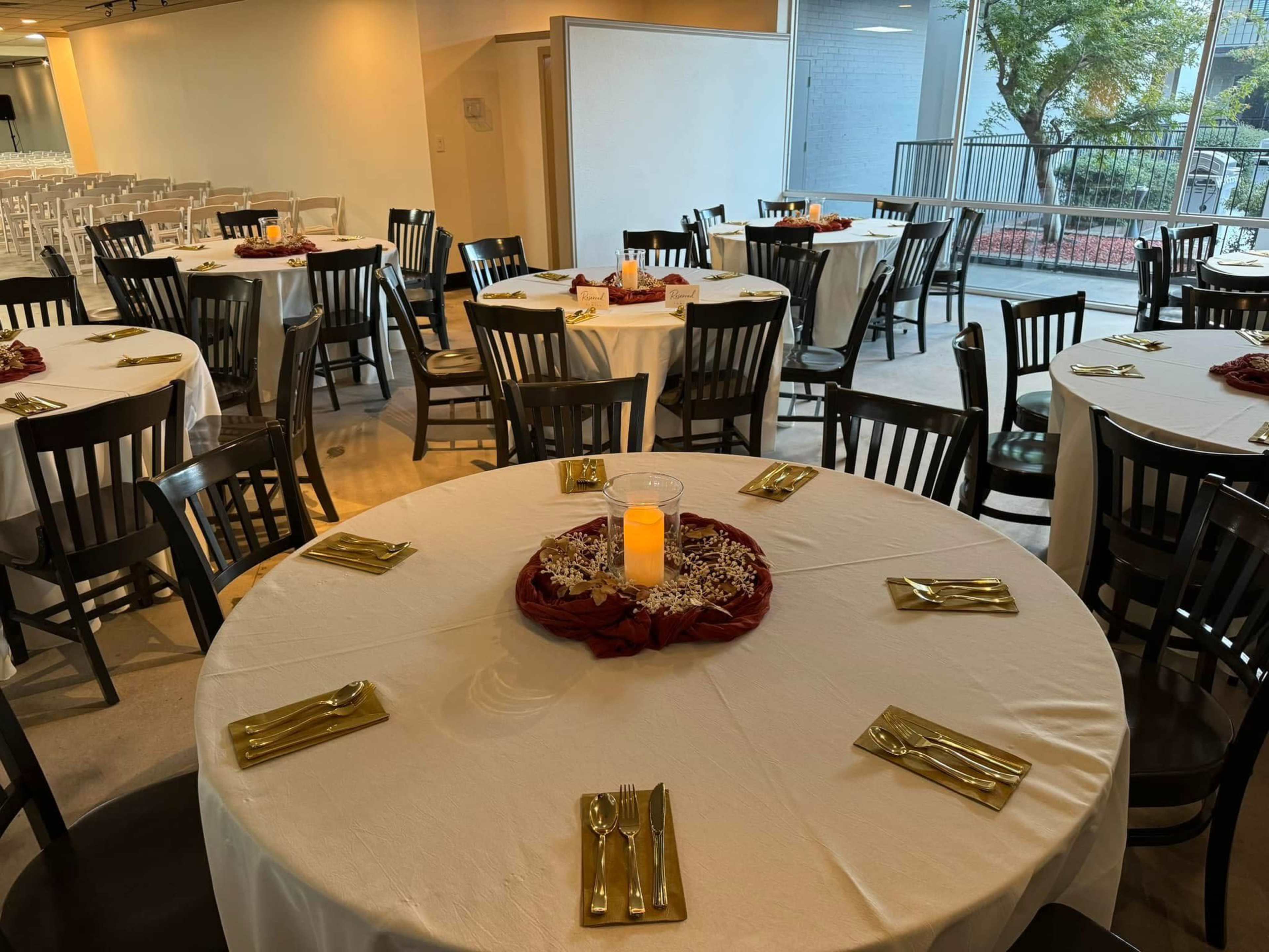 The image shows a dining area with several round tables covered in white tablecloths, each set with gold utensils and a centerpiece featuring a lit candle.
