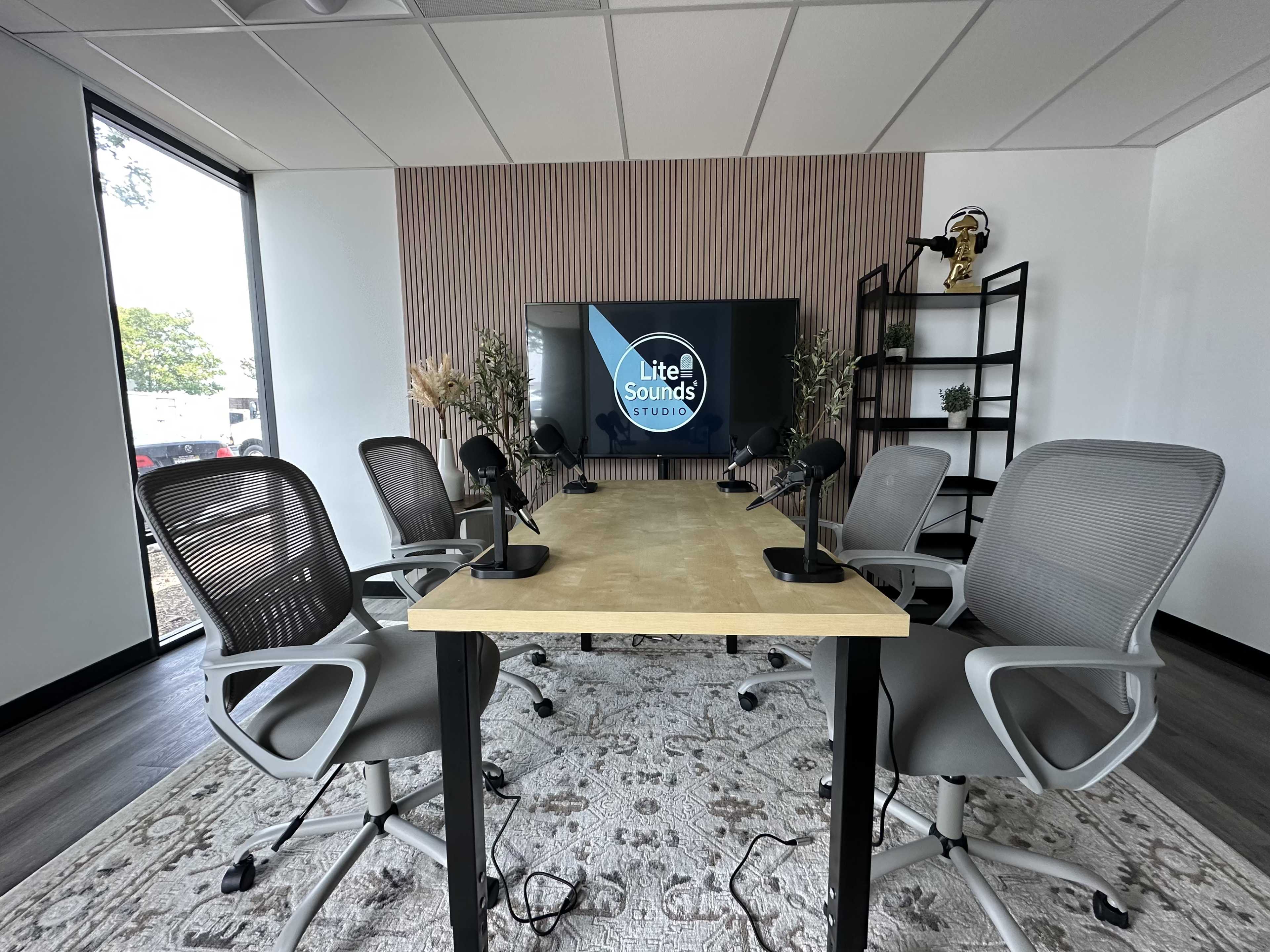 A modern podcast studio features a table with microphones and several ergonomic chairs, set against a backdrop of plants and a large screen displaying the "Lite Sounds Studio" logo.