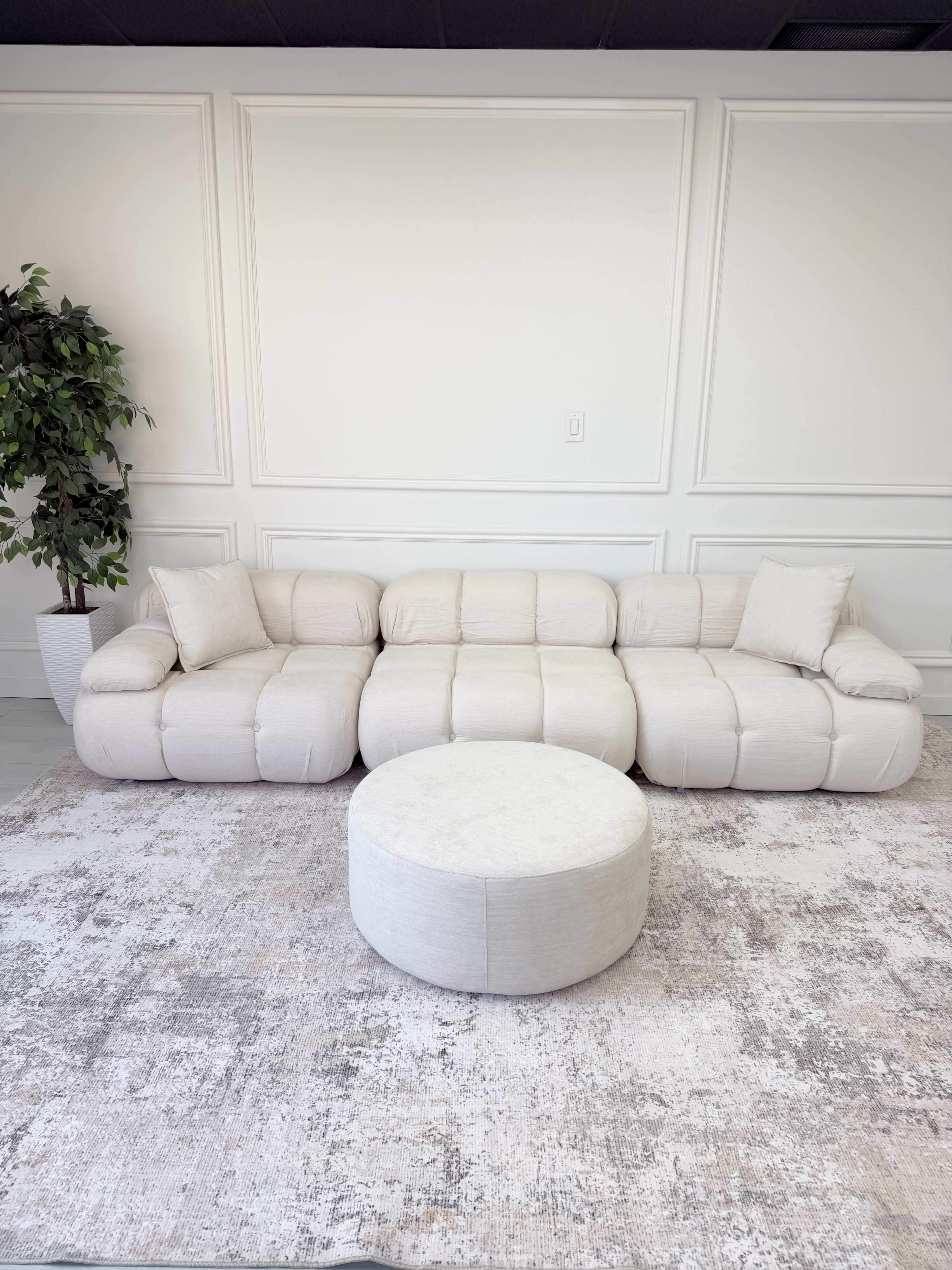 A light-colored, modern sectional sofa is positioned in a room with a round ottoman and a patterned area rug.