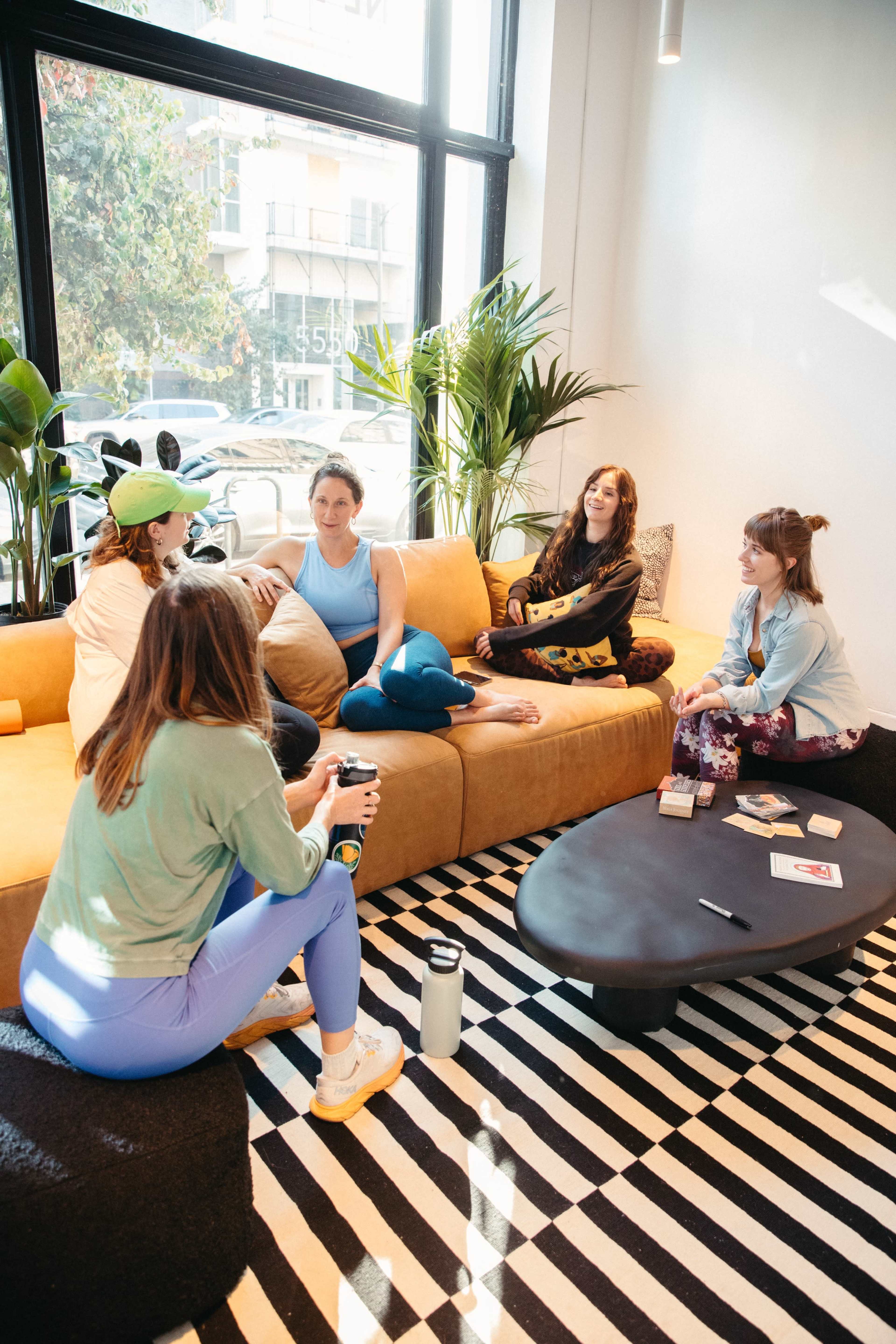 A group of five women are sitting around a black coffee table on a patterned rug in a brightly lit room with large windows and plants.