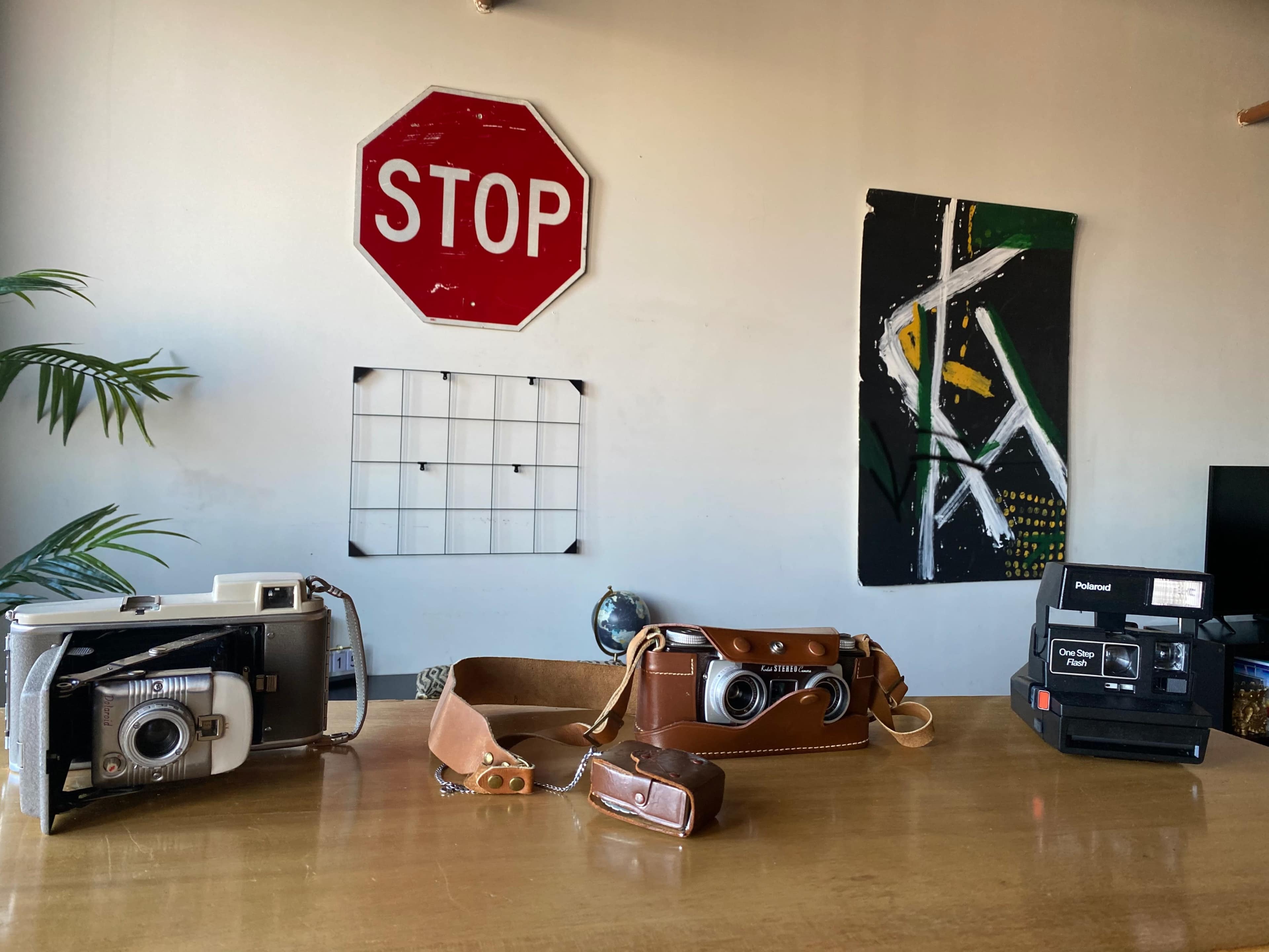 A wooden table with two vintage cameras, a leather camera bag, a globe, and a stop sign mounted on the wall behind them.