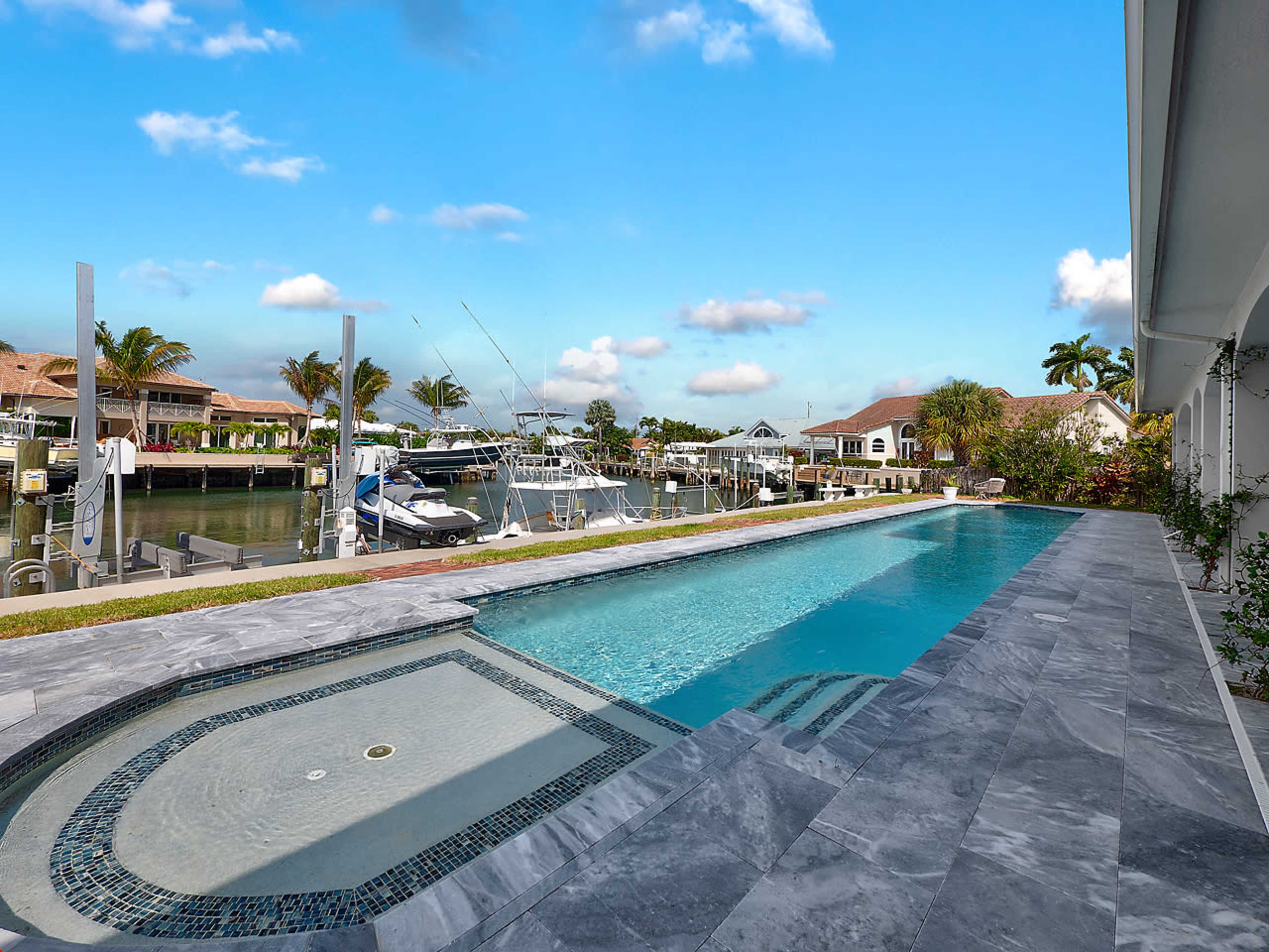 The image shows a modern outdoor pool next to a canal with several boats docked along the waterfront.