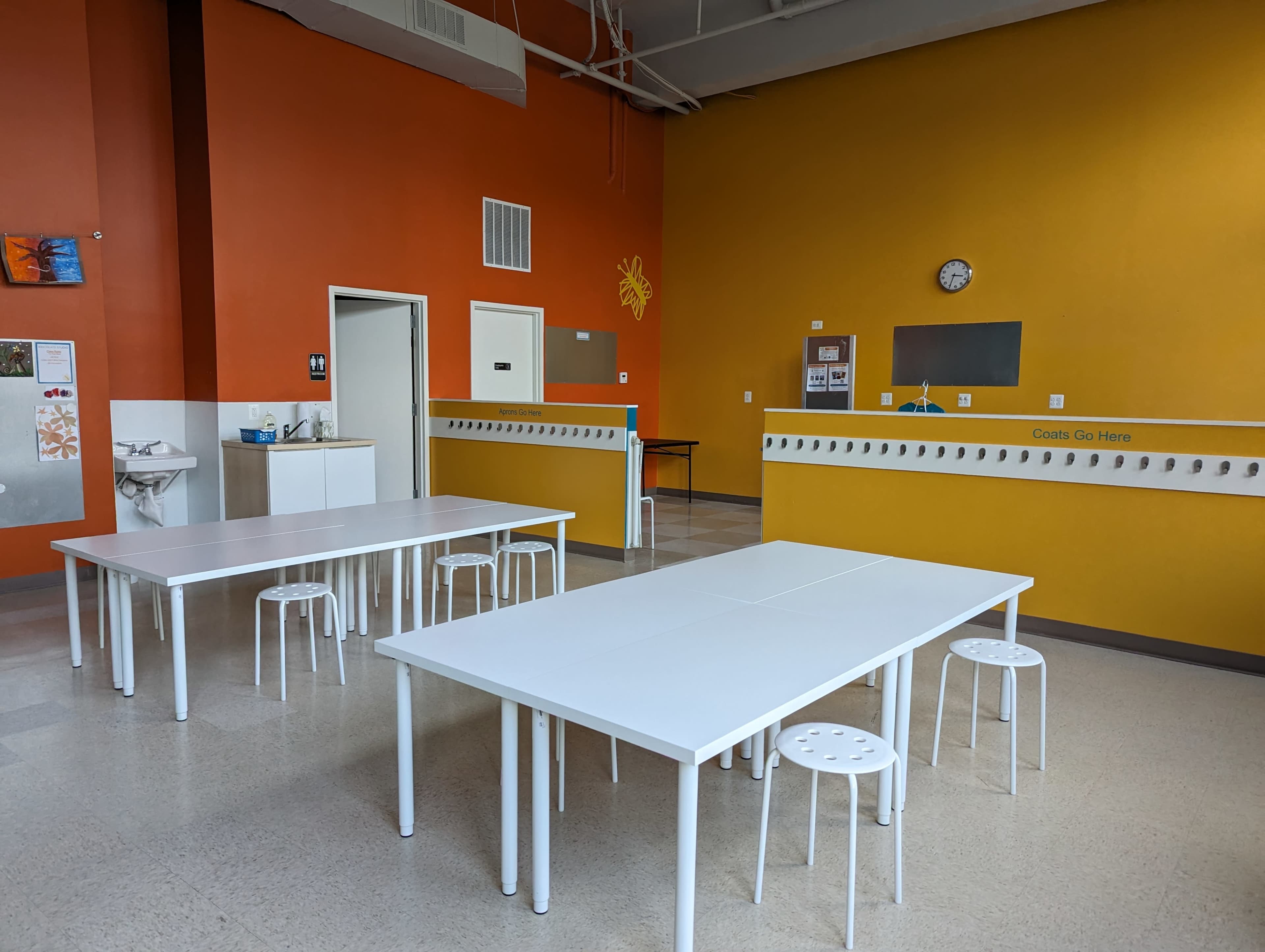 A spacious room with orange walls, two white tables, several white stools, and a sink area.
