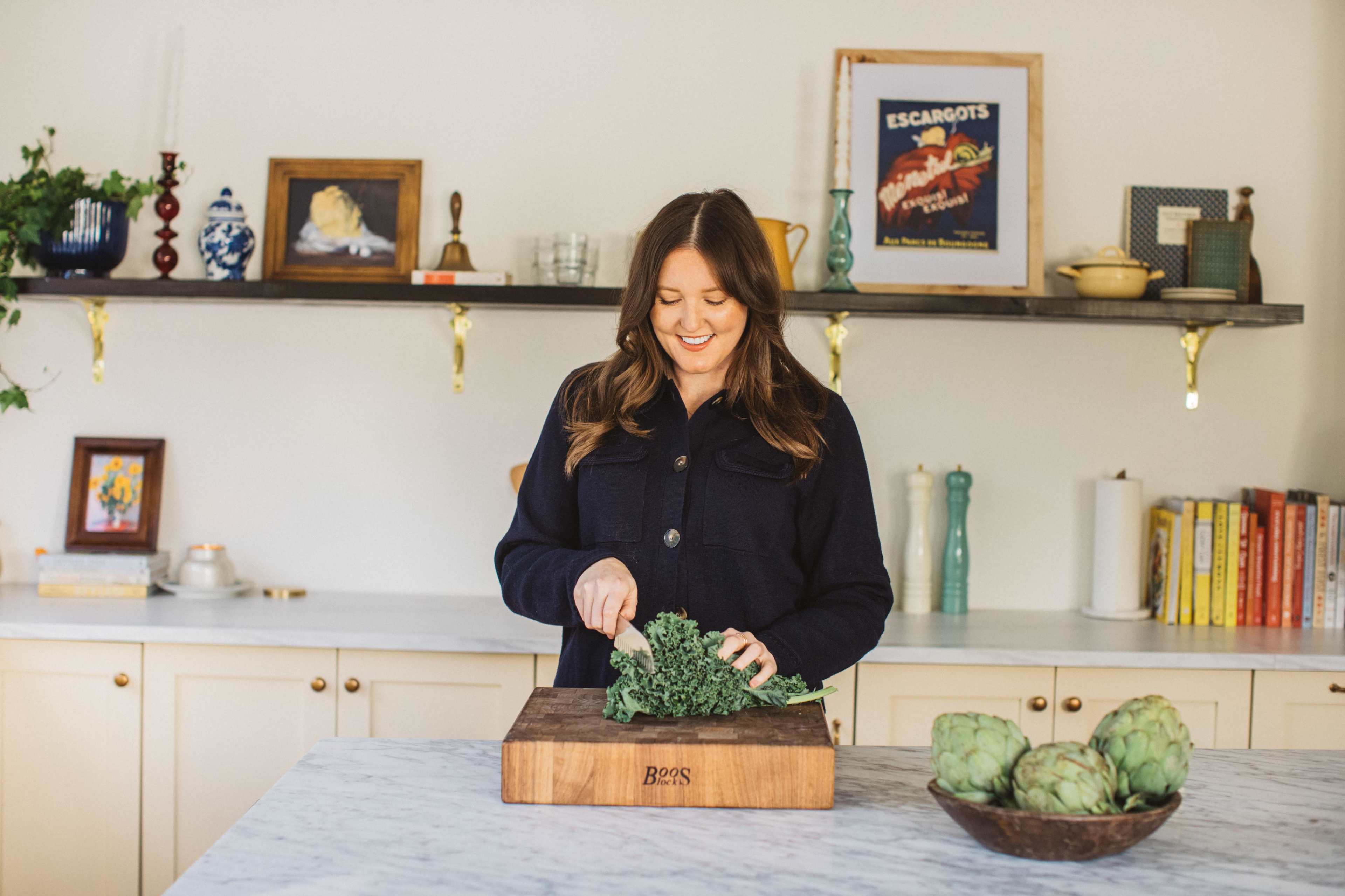 A woman is chopping kale on a wooden cutting board in a bright kitchen with decorative shelves in the background.