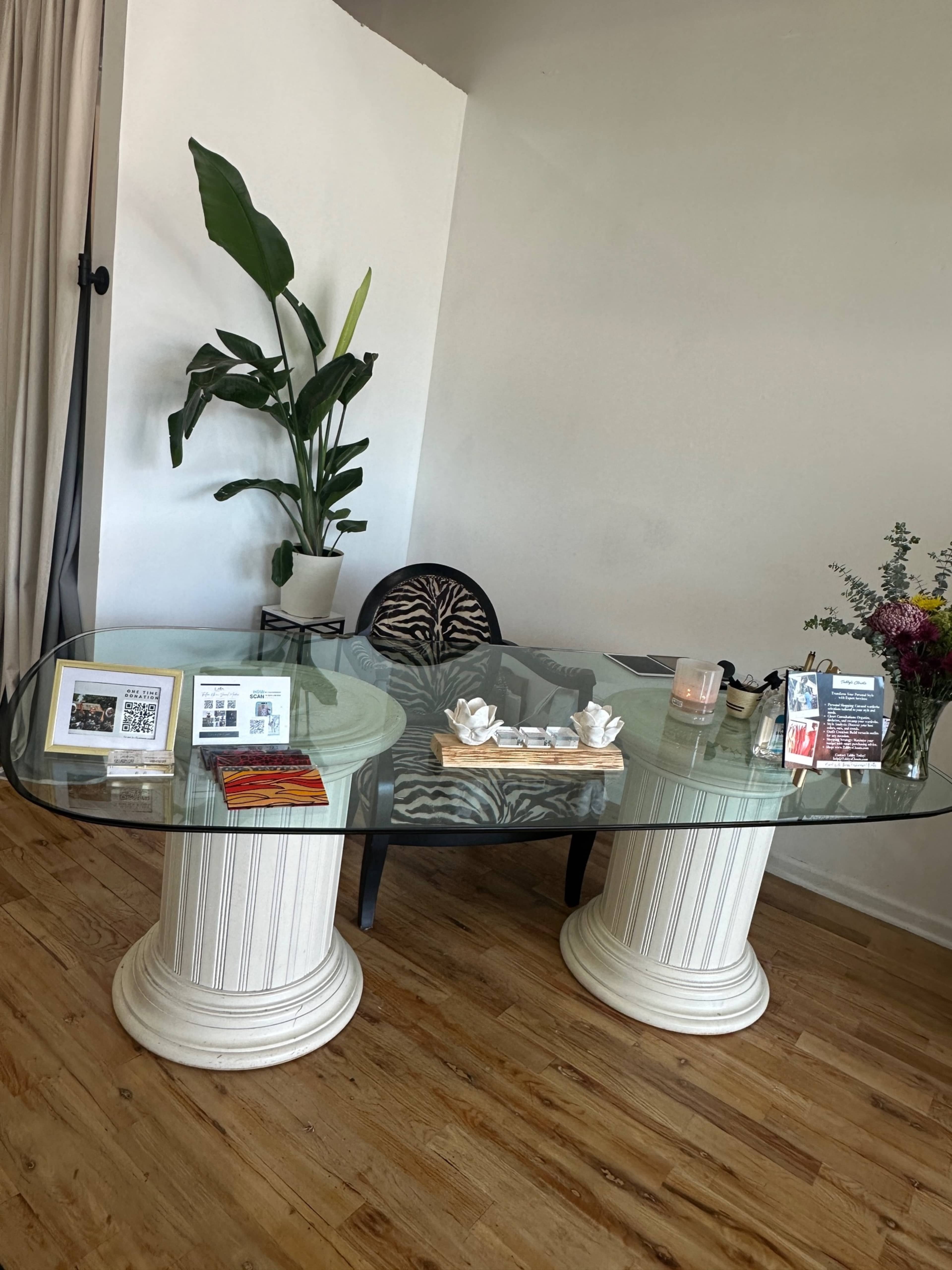 A modern desk with a glass top is set on two white columns, accompanied by a chair and a potted plant, with various brochures and a candle displayed on the surface.