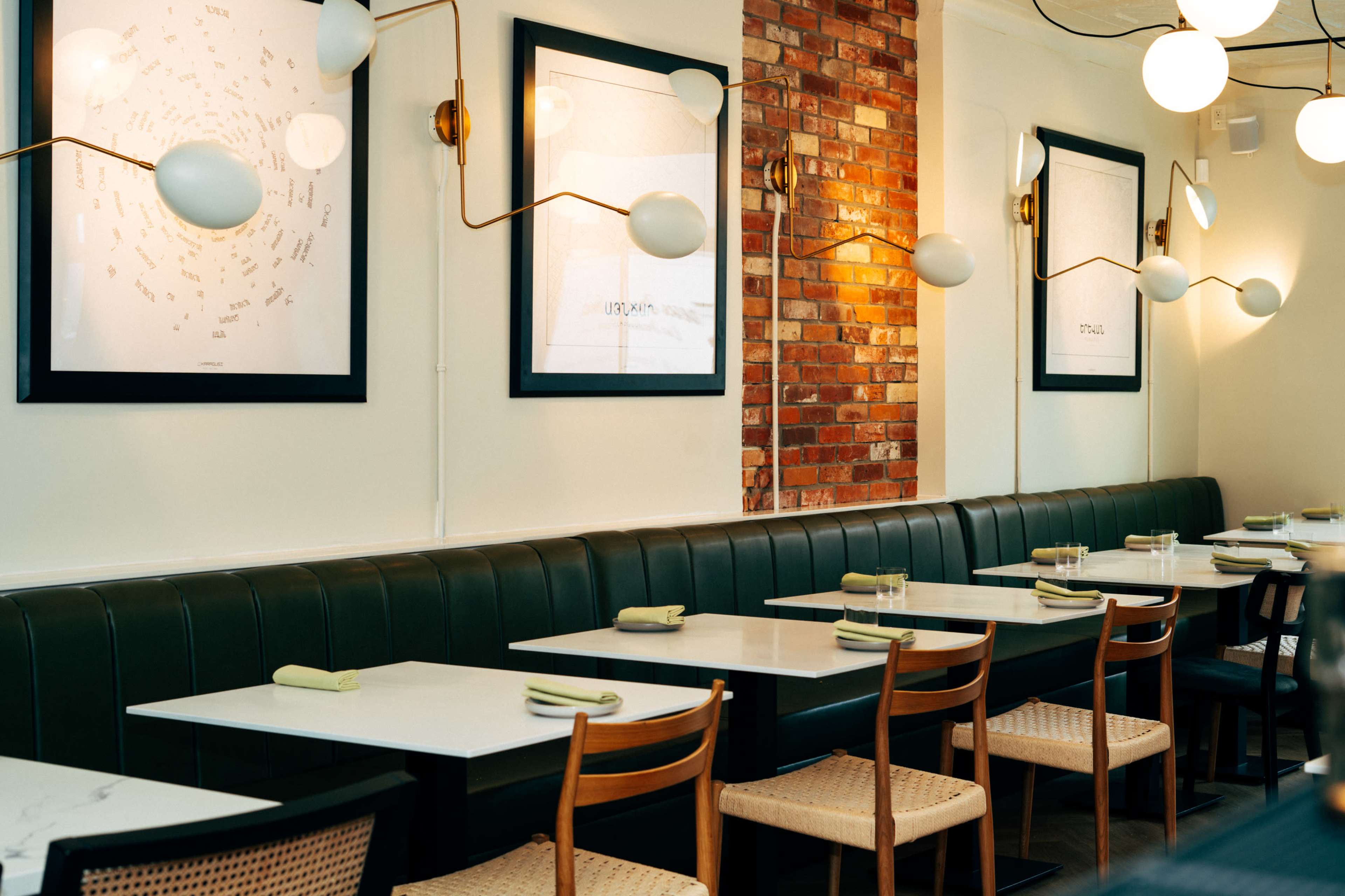 A dining area in a restaurant featuring green banquette seating, white tables, and artwork on the walls, illuminated by modern light fixtures.