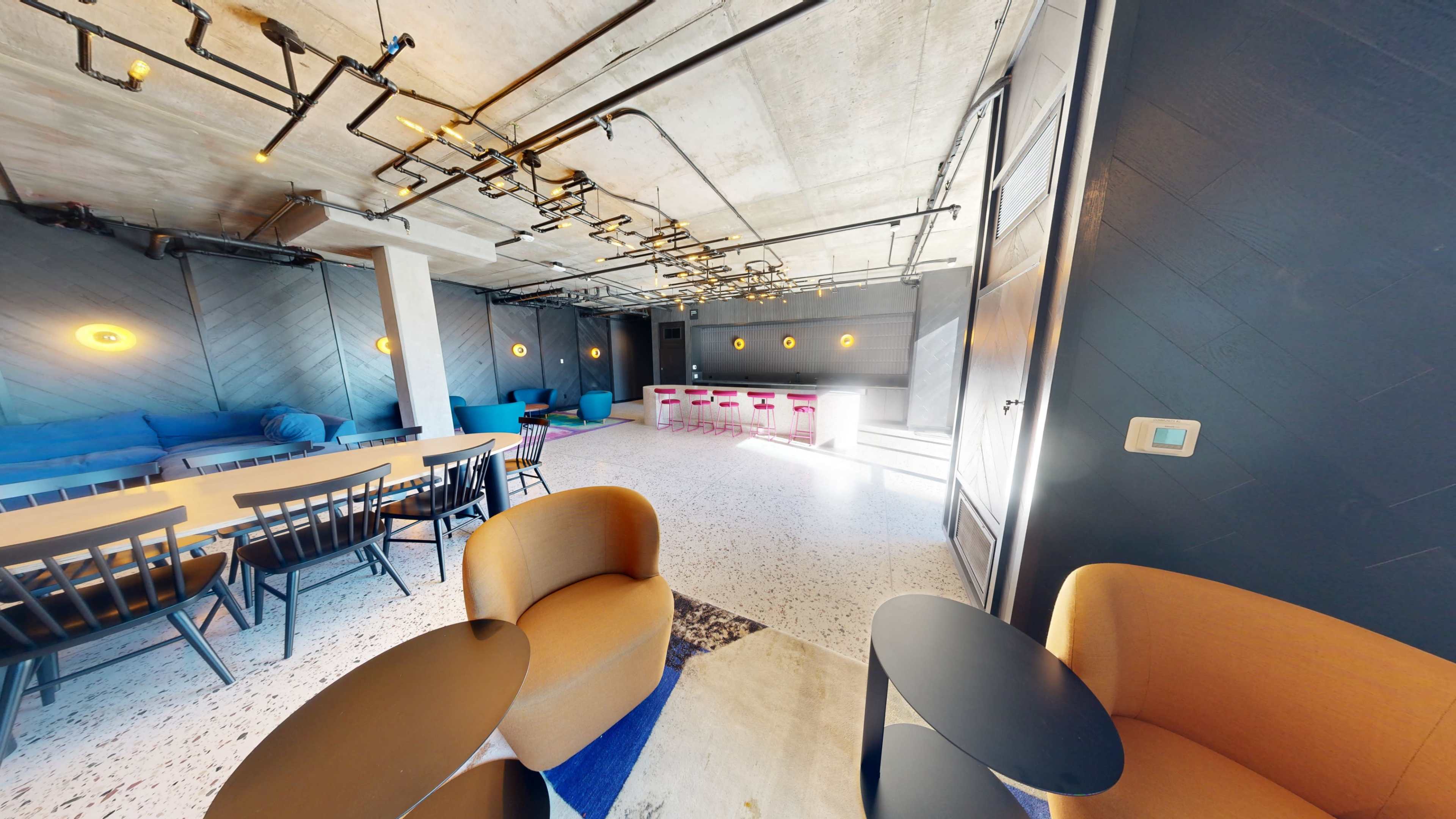 The image shows a modern indoor space with seating areas, a bar counter, and industrial-style lighting fixtures.