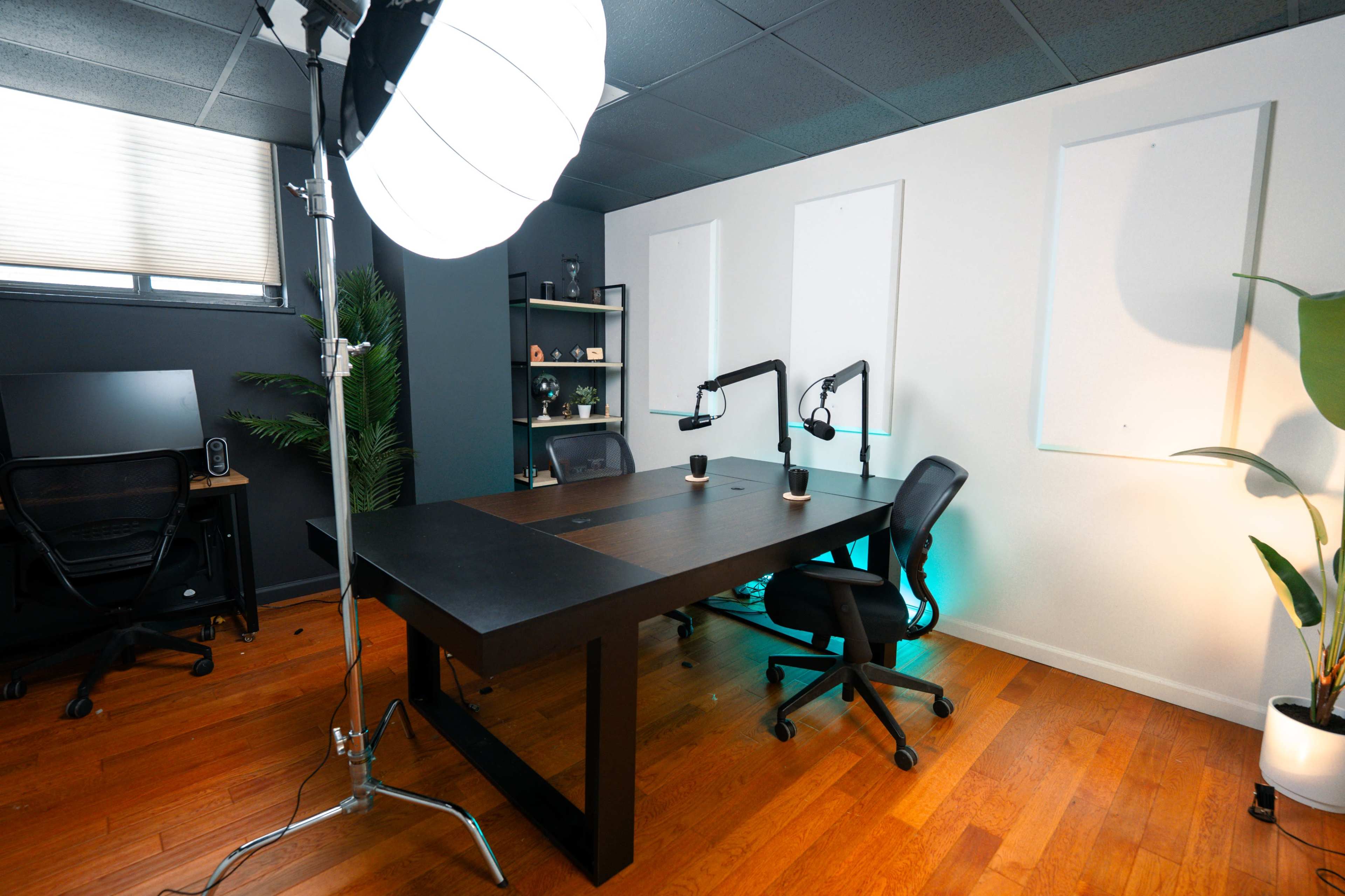 The image shows a modern office space with a large black table, two office chairs, a computer setup, and a softbox light, all against a backdrop of grey walls and wooden flooring.