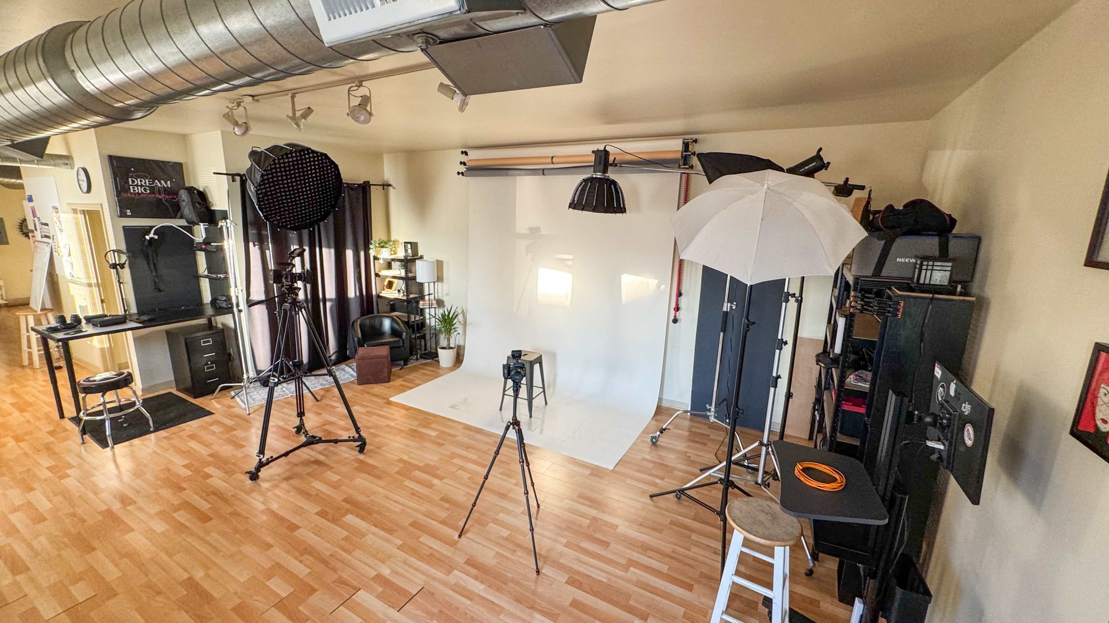 The image shows a photography studio equipped with various lighting setups, backdrops, and props arranged in a spacious area with wooden flooring.