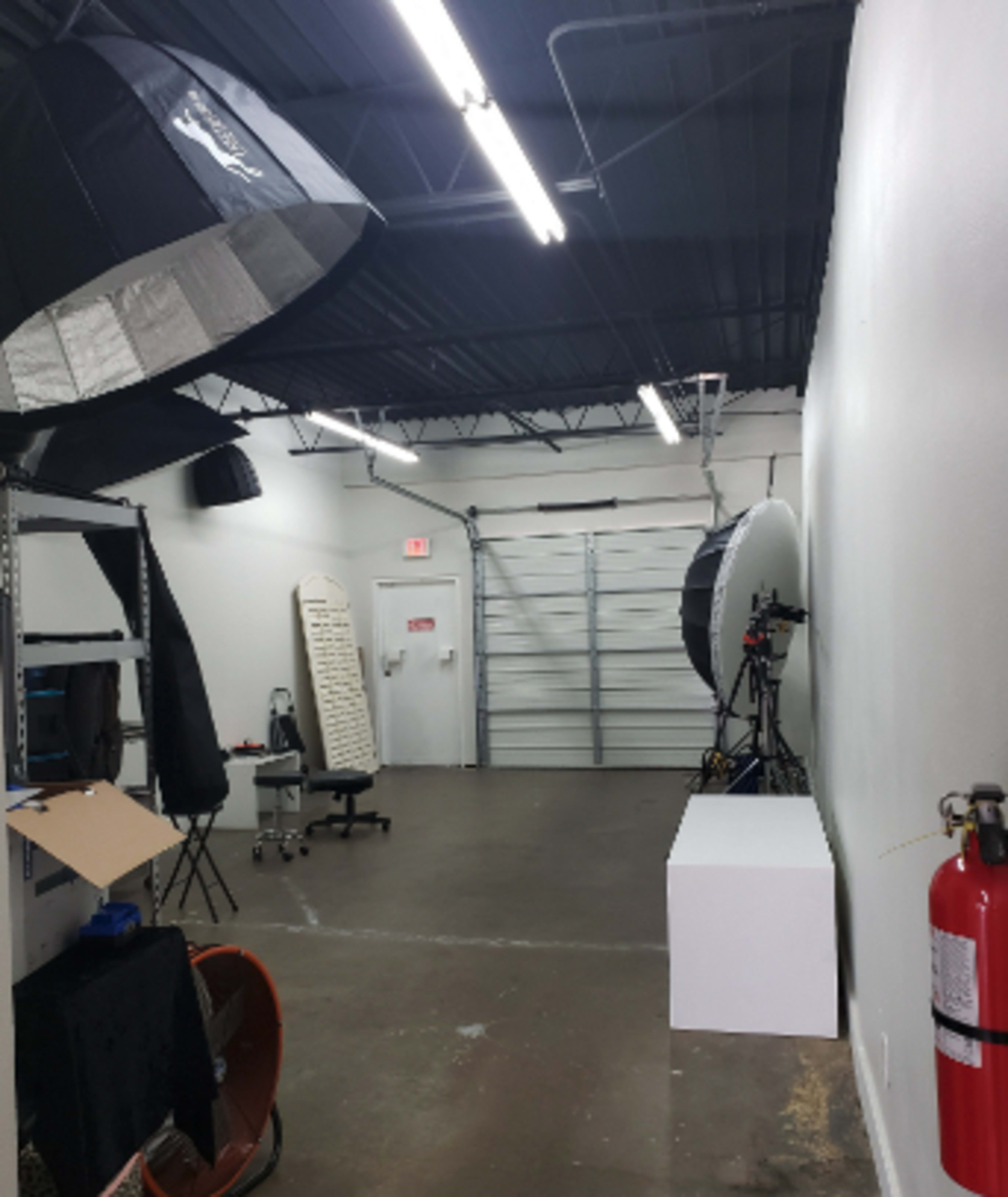 The image shows a photography studio with lighting equipment, a white backdrop, and a garage door at one end.