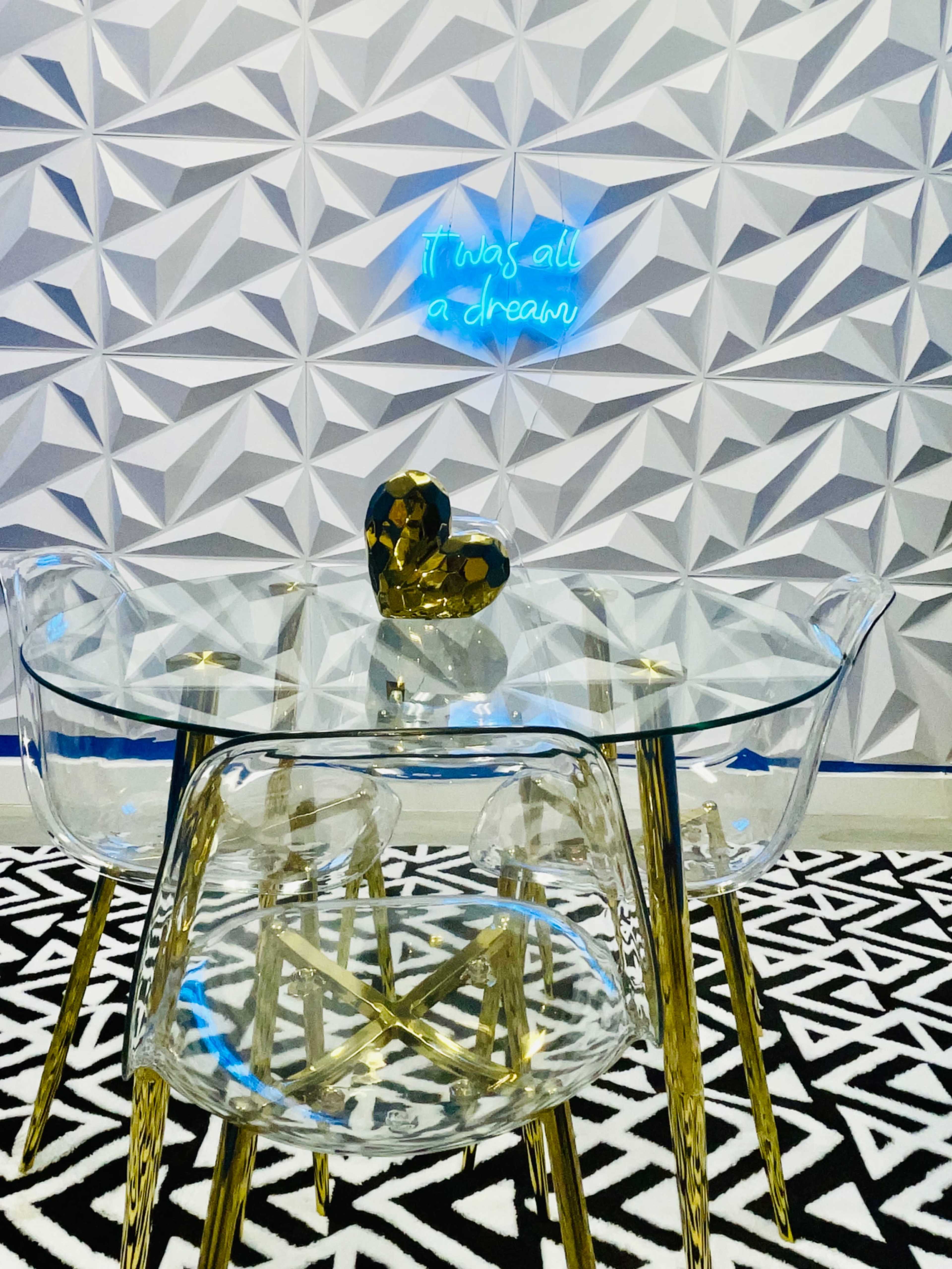 A glass table with gold legs and a heart-shaped decor piece sits on a patterned black and white rug, beneath a geometric wall featuring a neon blue sign that reads, "It was all a dream."