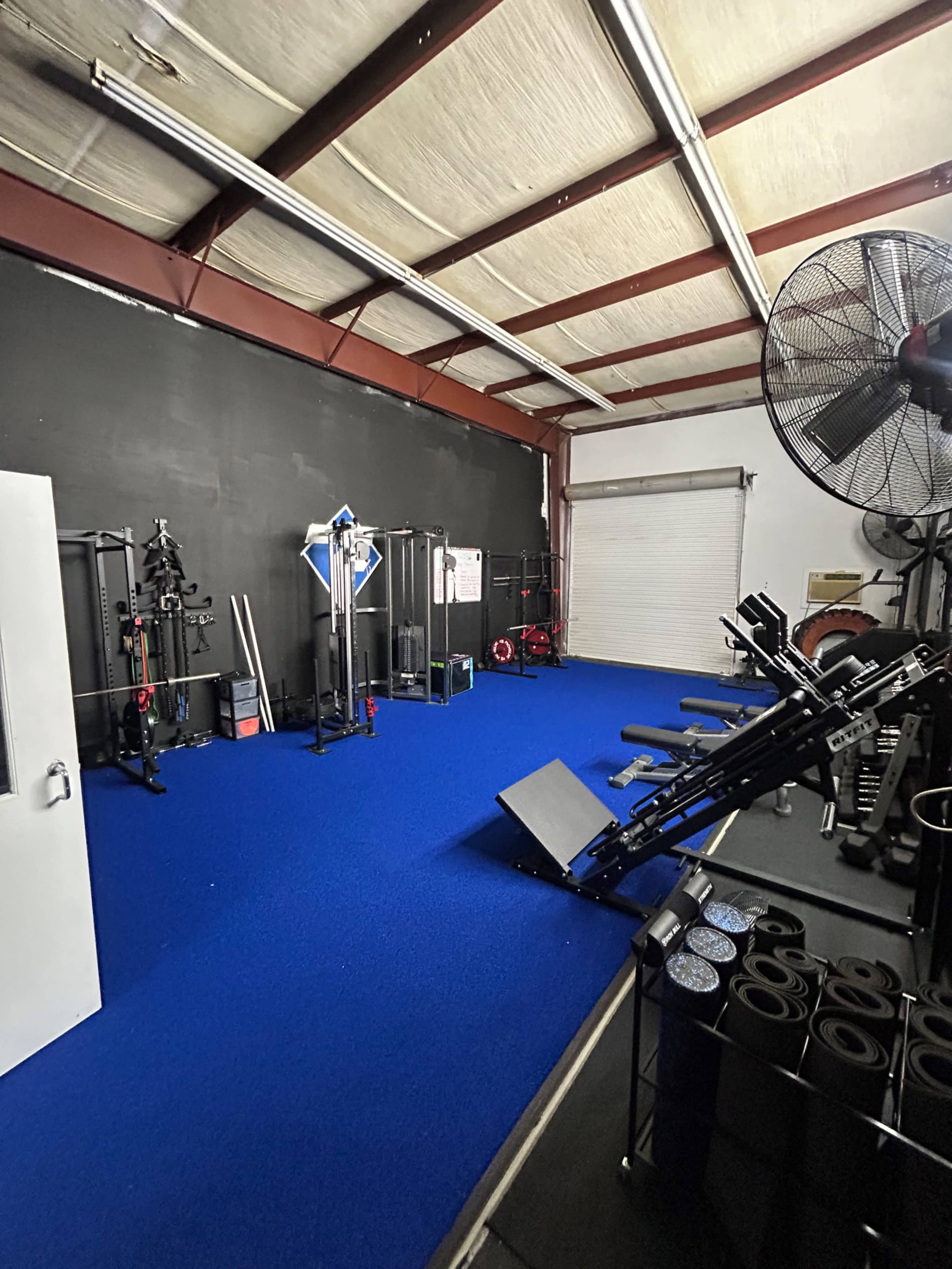 The image shows a gym space with blue synthetic flooring, various fitness equipment, and a large overhead fan.