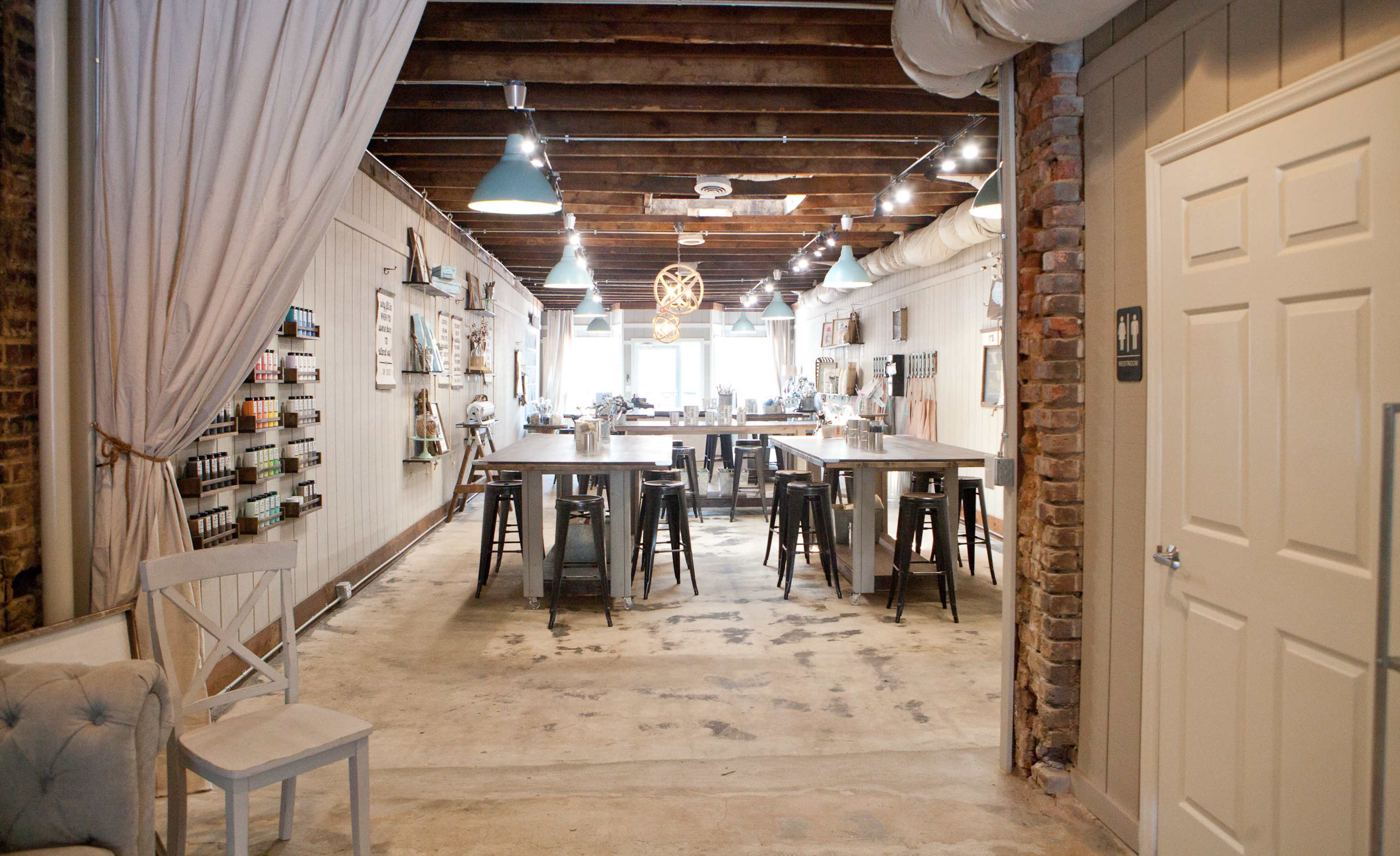 A spacious interior of a creative workspace with tables, stools, and art supplies arranged along the walls under exposed wooden beams.