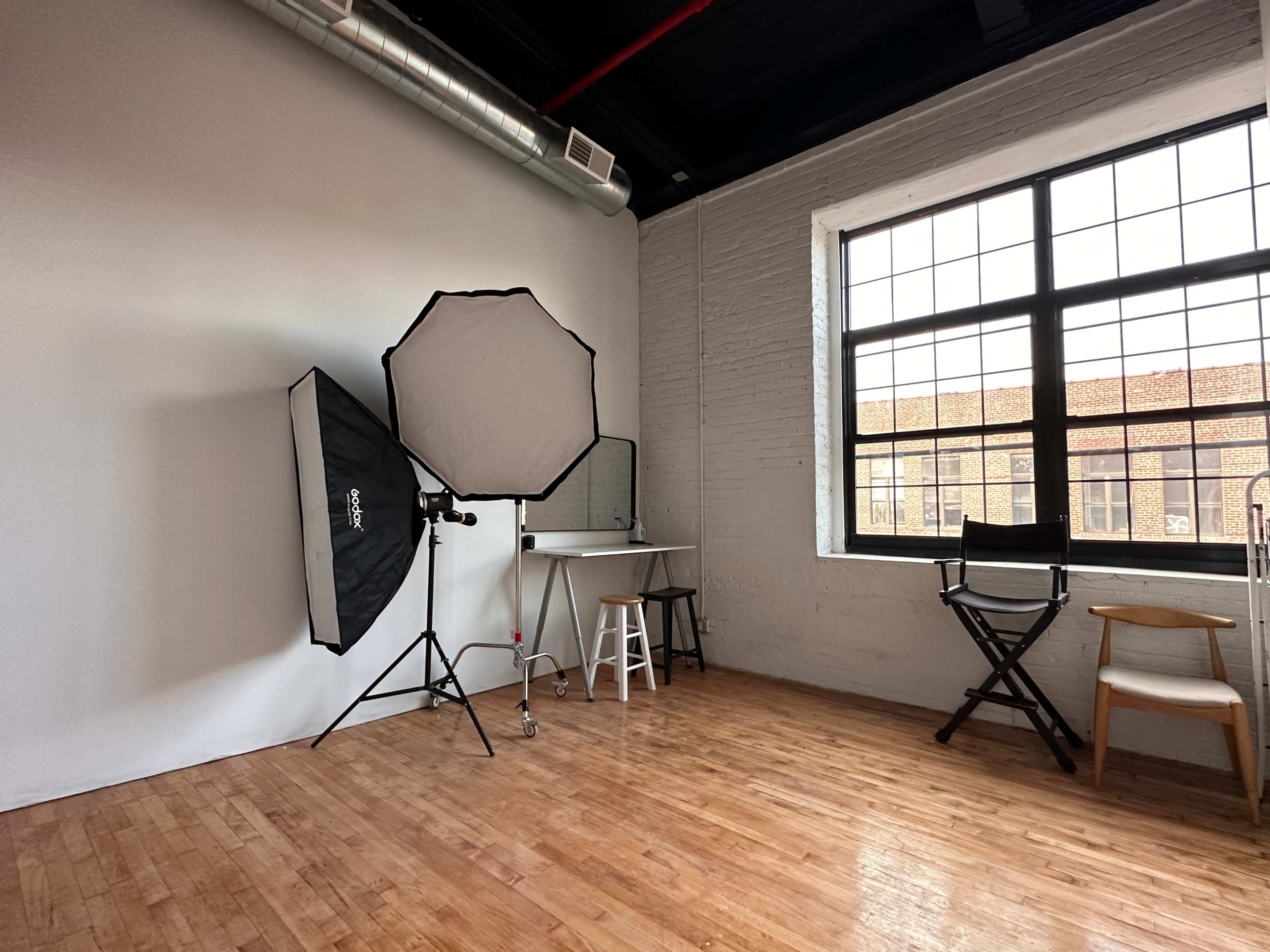 A photography studio with a large softbox light, a mirror on a table, and director's chairs situated against a brick wall.