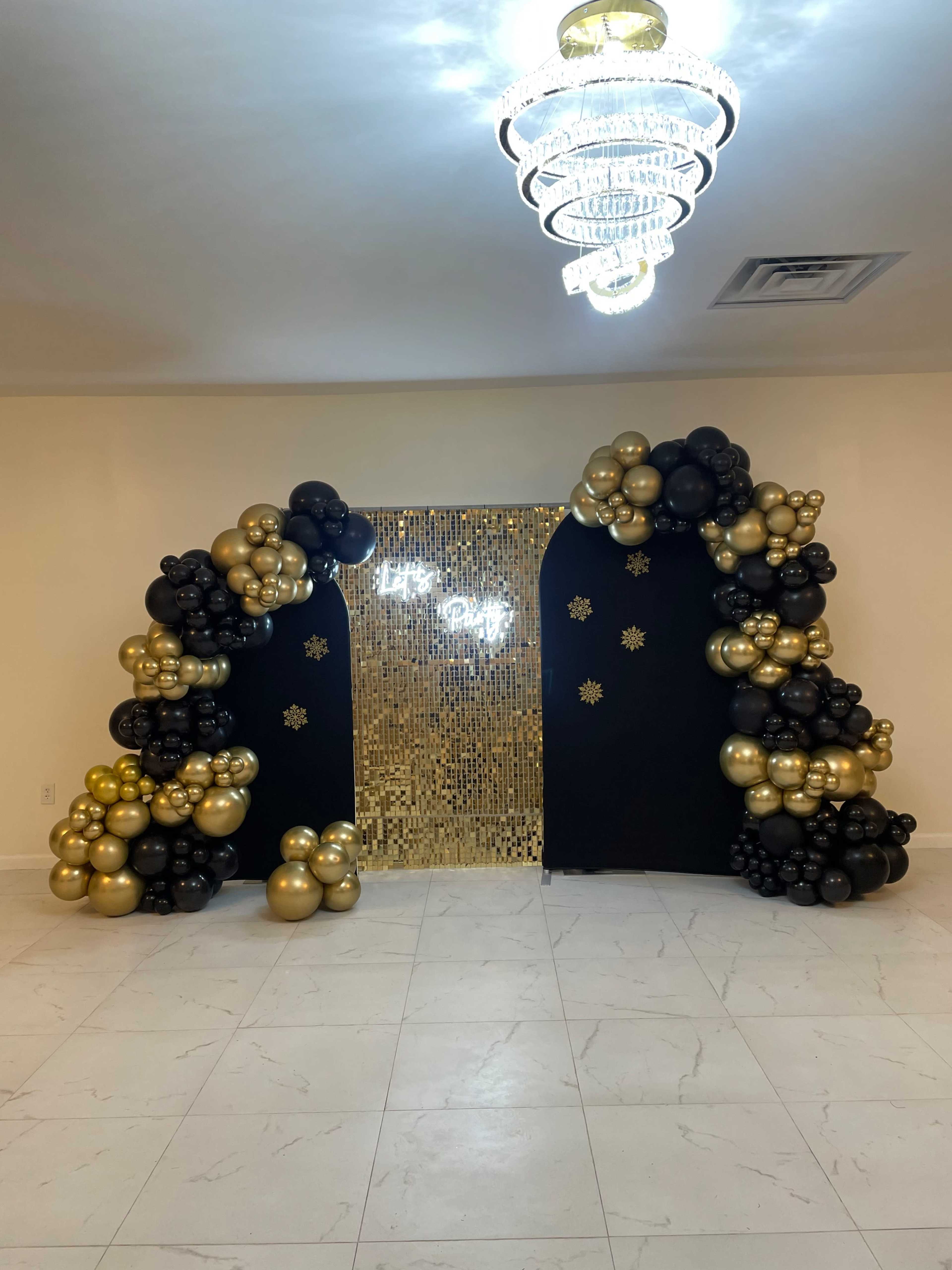 The image shows a festive decor setup with a backdrop of black and gold balloons surrounding a glittering gold wall and illuminated text reading "Let's Party."