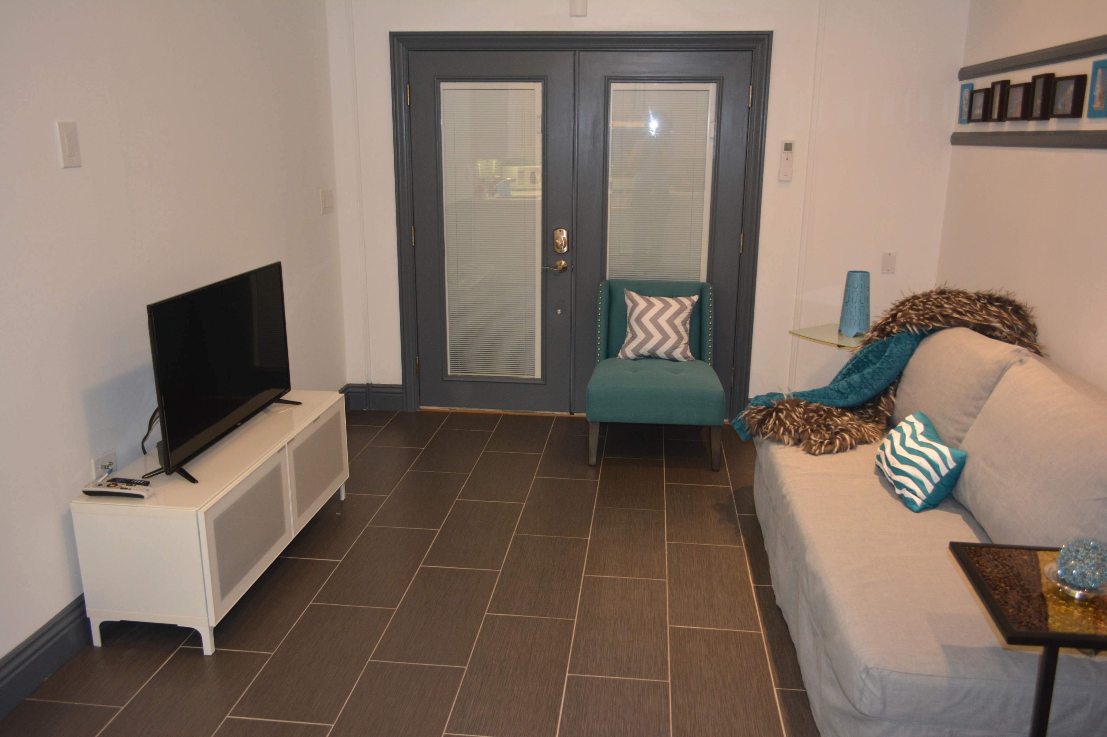 A modern living room features a gray sofa, a turquoise chair, a television on a white stand, and a glass table with decorative items.