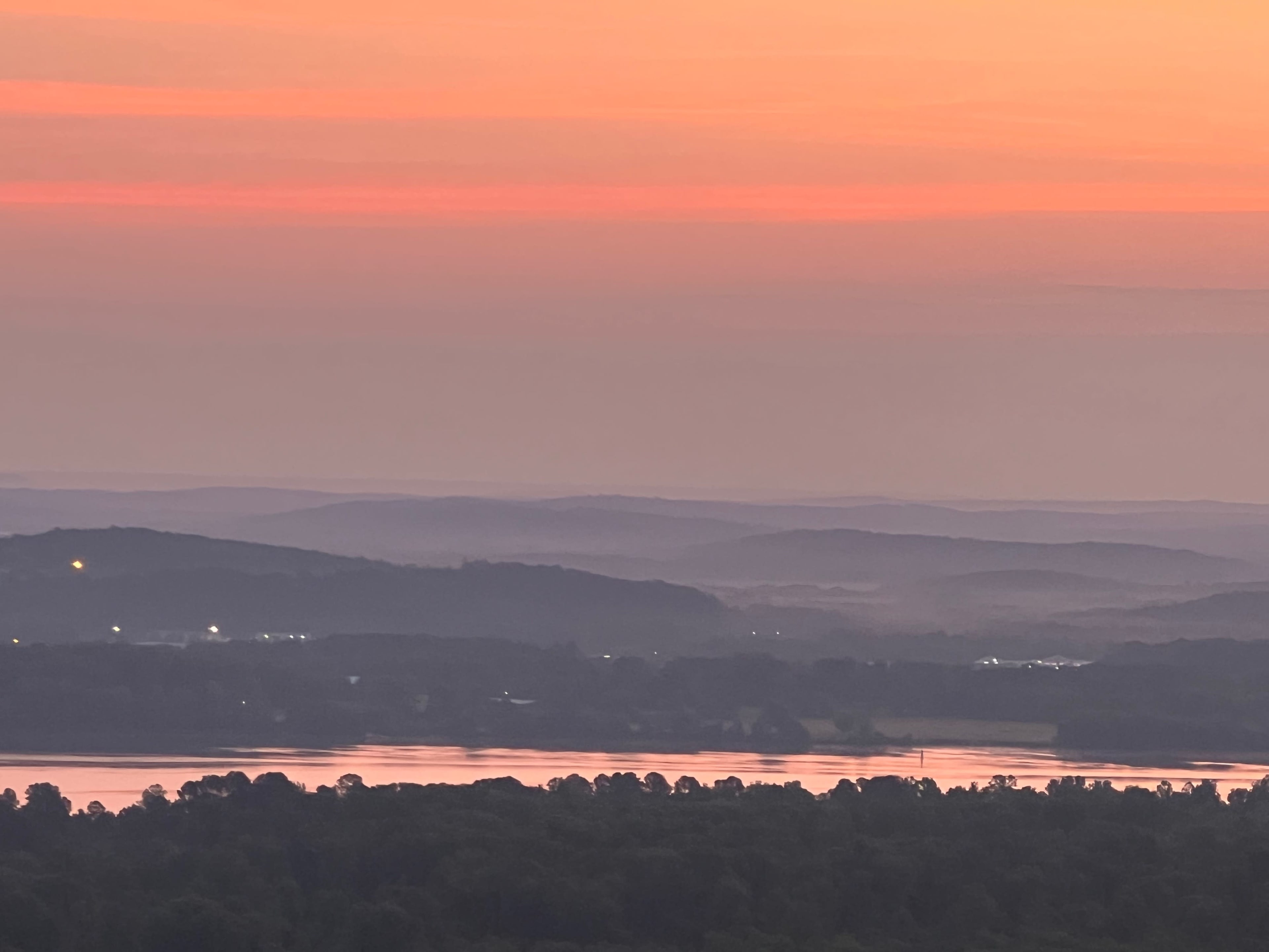 A tranquil landscape at dusk, with layered hills and a river reflecting soft hues of orange and pink in the sky.