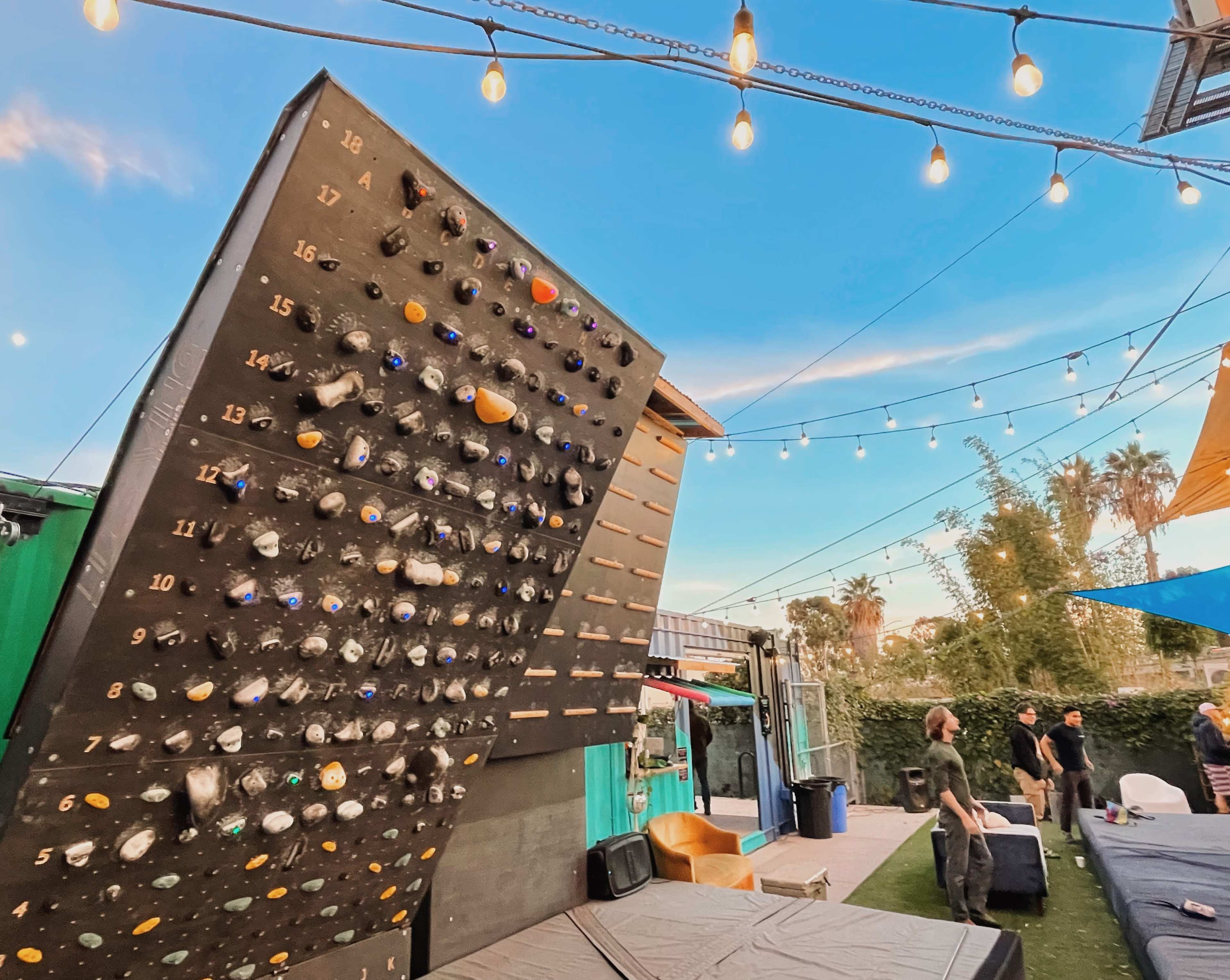 A climbing wall with various handholds stands under string lights in an outdoor recreational area.