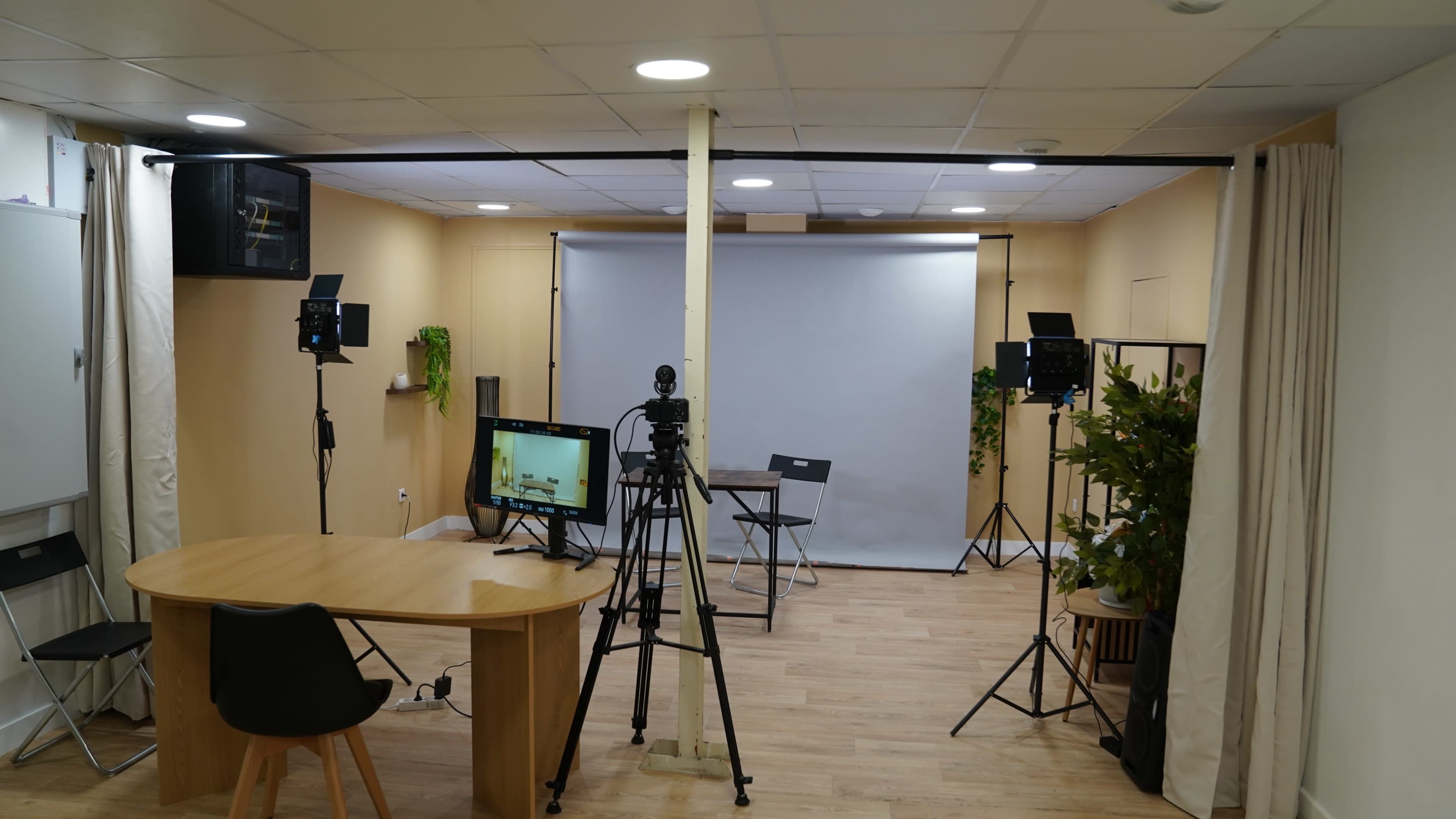 The image shows a video production studio with a large backdrop, camera equipment on tripods, a computer monitor, a table, and chairs.