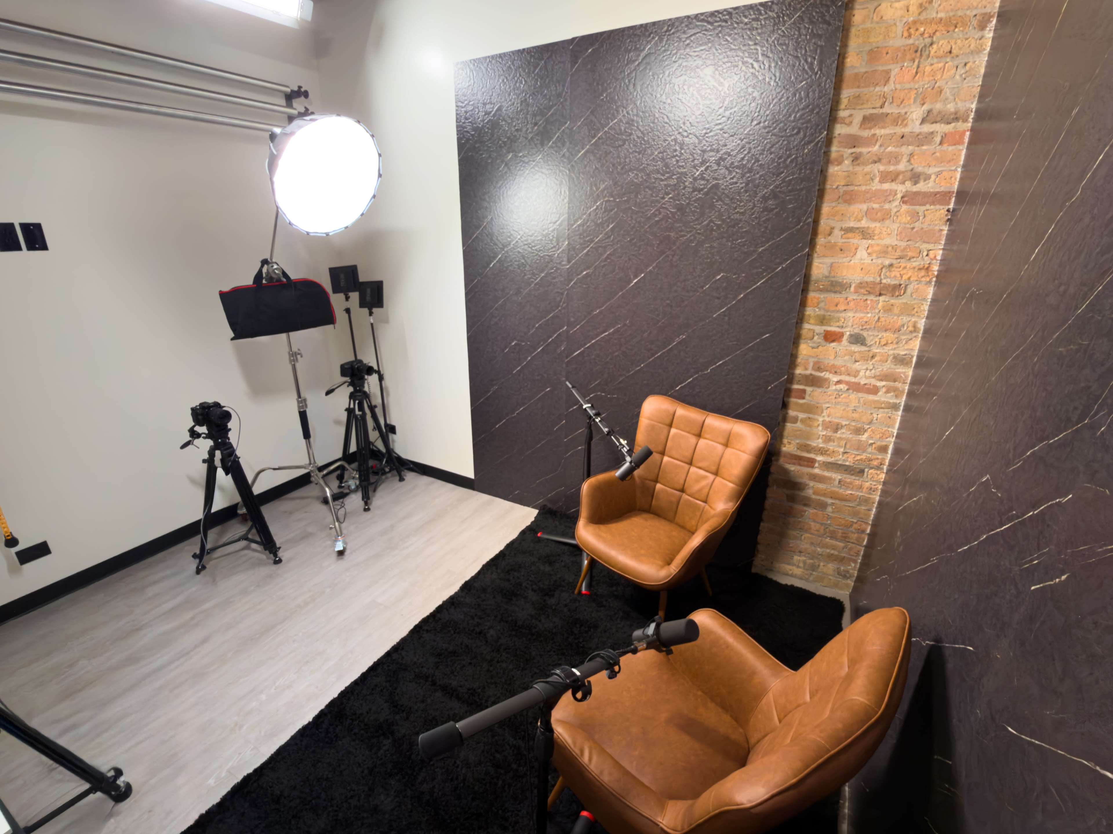 The image shows a recording studio setup featuring two brown chairs, a dark textured wall, and several cameras and lighting equipment arranged on the floor.