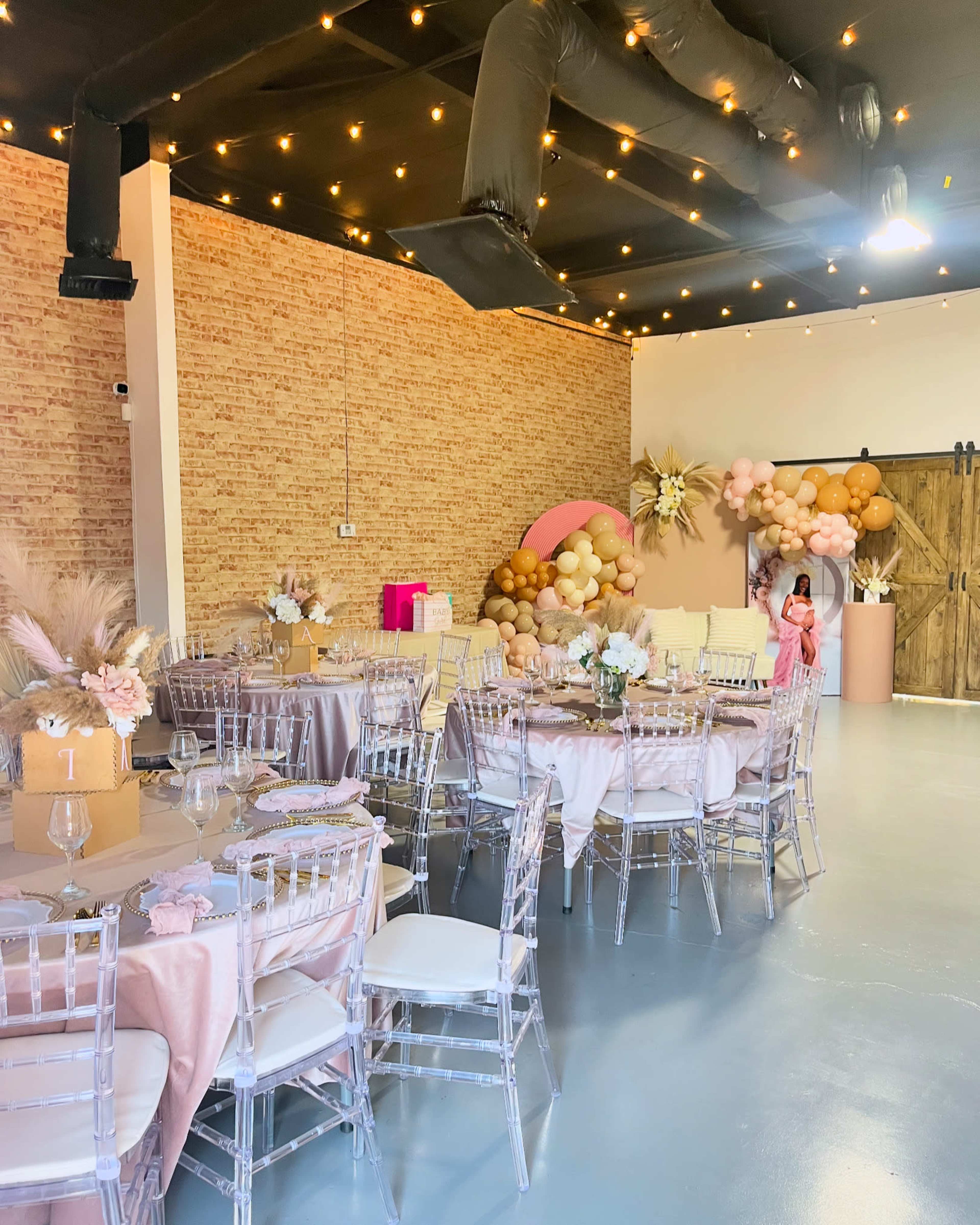 A decorated event space with round tables, transparent chairs, and a backdrop featuring balloons and floral arrangements.